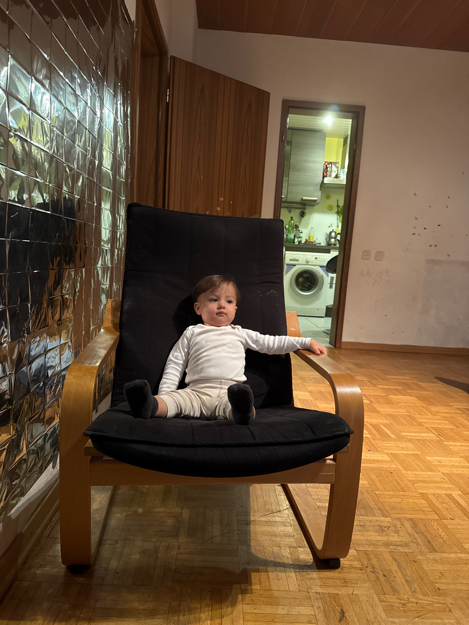 In Überherrn, Germany, a young child sits in a black recliner chair with a wooden frame. The child, wearing a white long-sleeved shirt and light-colored pants, is positioned centrally within the frame. The child is facing slightly to the right, with their right arm extended and resting on the armrest. The background reveals a partially visible kitchen with a washing machine and cabinets, visible through an open doorway. To the left, a wall is covered with a reflective, square-tiled material that creates a mosaic effect. The floor is a light-colored parquet. The lighting suggests an indoor setting, possibly in the evening or with artificial lighting.