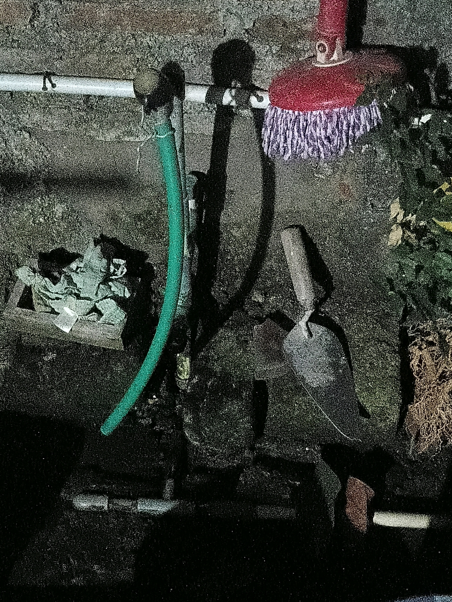 The image depicts an outdoor scene, possibly a backyard or a utility area, with a rough, textured wall in the background. A white pipe runs horizontally across the upper left of the frame, supported by brackets. Attached to this pipe is a green garden hose, coiled loosely. To the right of the hose, a red mop head with white and purple bristles hangs from a handle, casting a shadow on the wall. In the lower center, a trowel with a dark metal head and a wooden handle rests on the ground. To the left of the trowel, a wooden box contains what appears to be crumpled green fabric or paper. Further down, a section of PVC pipe is visible on the ground. The ground itself is uneven and covered with dark material, suggesting dirt or shadow. The scene appears to be located in Cikal, Indonesia, although no specific landmarks or identifying features within the city are visible in the image.