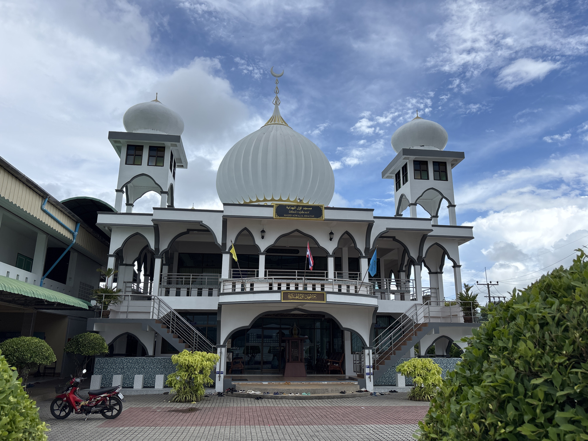 The image shows the exterior of Masjid Aow Pree Hidaya, a mosque located in Rawai, Thailand. The mosque is a two-story white building with a prominent central dome topped with a crescent moon. Two smaller domes are situated atop two flanking minarets. Arched windows and doorways are visible throughout the structure, with grey trim accentuating the white facade. A balcony with a railing extends across the second floor, adorned with flags of Thailand, and two other unidentified flags. The entrance to the mosque is at ground level, with stairs leading up to the balcony. In front of the mosque, a red motorcycle is parked on a red and grey tiled courtyard. Lush green bushes and small trees are planted around the building, and a section of an adjacent building with a green roof is visible to the left. The sky is partly cloudy.