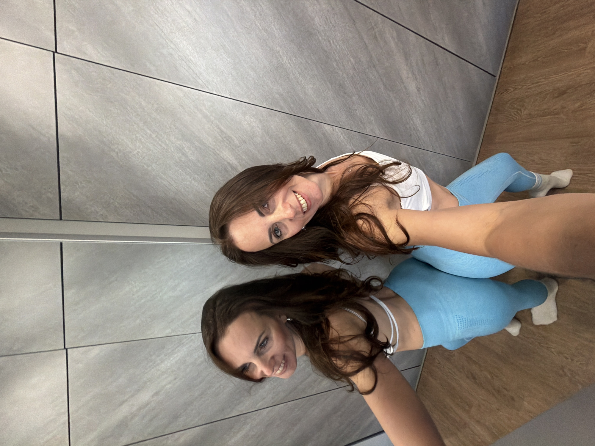 An indoor selfie captures a smiling young woman with long, dark brown hair. She is dressed in a white sports bra or top and light blue athletic leggings, complemented by white socks. The woman holds the camera to capture her reflection in a large mirror, which is prominently visible directly below her in the frame. Her reflection mirrors her pose and cheerful expression.
The background setting comprises a clean, light gray tiled or paneled wall with dark grout lines, suggesting a modern aesthetic. A wooden floor is visible at the bottom of the image, where the woman's legs and the lower part of her reflection are positioned. The overall scene conveys a bright, indoor environment, likely a gym, fitting room, or a contemporary private space. The woman's relaxed posture and smile indicate a casual moment, possibly after a workout or during a style check. No text is visible in the image. The location is Birobidzhan, Russia.