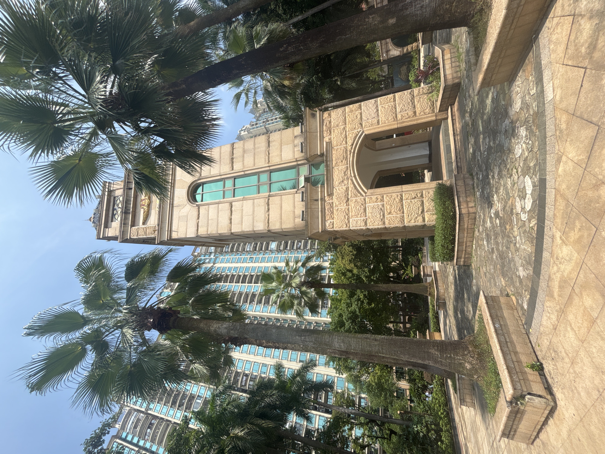 In Nanhai District, China, a tall, stone clock tower stands prominently in a courtyard. The tower features a large arched entrance at its base and a long, narrow, green-tinted window on its upper section. The clock face is partially visible. Several mature palm trees surround the tower, their fronds reaching towards a clear blue sky. In the background, a modern, high-rise apartment building with many balconies and light blue-tinted windows rises above the trees. The ground in the foreground is paved with stone tiles of various shapes and colors, and several raised planters containing greenery are visible. The scene suggests a well-maintained outdoor space within a developed urban area.