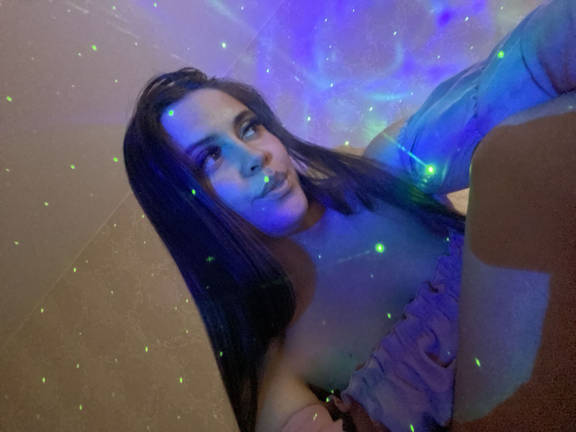 The image shows a young woman with long, dark hair, looking up and to the right, with her head tilted. She is wearing a light-colored, off-the-shoulder top with ruffles. The background is dimly lit and features a projection of green and purple lights, creating a starry or nebulous effect. The lighting casts reflections and shadows on the woman's skin and clothing. The scene appears to be indoors, with a textured wall visible in the upper left corner. The overall atmosphere is intimate and relaxed, possibly in a bedroom or living space. There is no specific information in the image that would allow for a determination of its location within Chaikovsky, Russia.