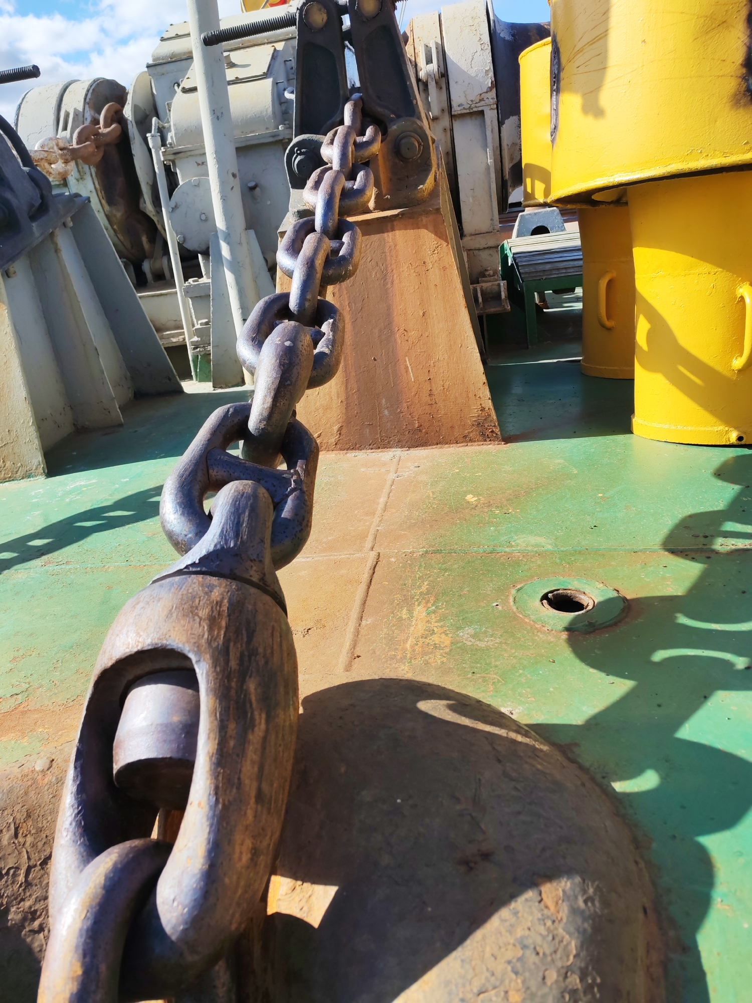 A close-up shot depicts a large, weathered metal chain, likely an anchor chain, resting on the green deck of a ship. The chain links are substantial and show signs of rust and wear, suggesting age and heavy use. In the background, machinery and structures of the ship are visible, painted in yellow and gray. A small table can also be seen.
There is no indication of Rybinsk, Russia in this image.