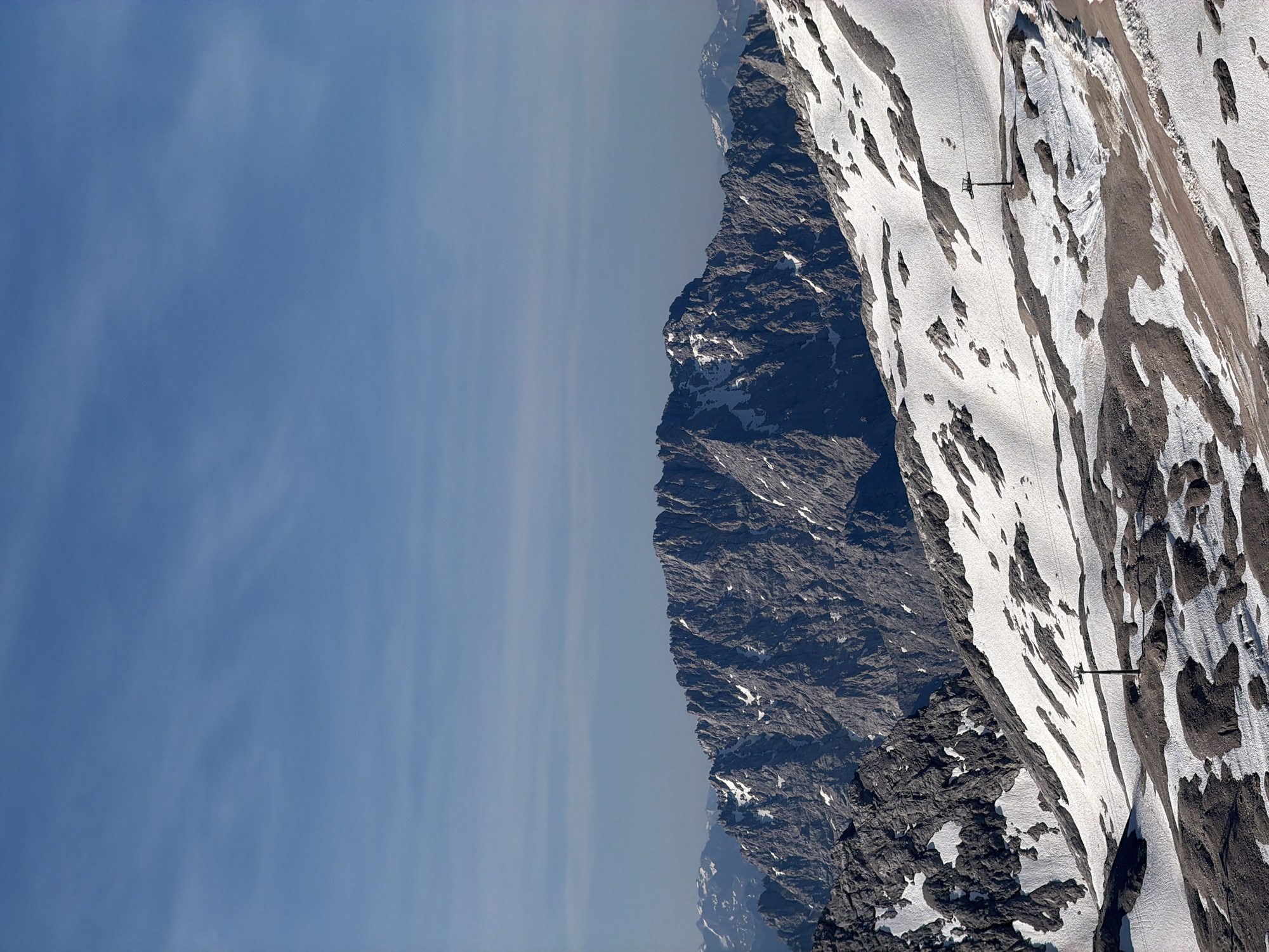 This is a photograph of a mountainous landscape with snow on the ground. In the foreground, there is a snowy slope with scattered rocks and a single ski lift pole. In the midground, a large, rugged mountain range with patches of snow dominates the scene. In the background, distant mountains are visible under a clear blue sky with thin, wispy clouds. The image appears to be taken from a high vantage point, likely a mountaintop or a cable car. The location is likely the Zugspitze, the highest peak in Germany, which is near Garmisch-Partenkirchen. There are no people or animals visible in the image. The overall scene is one of natural grandeur and alpine beauty.