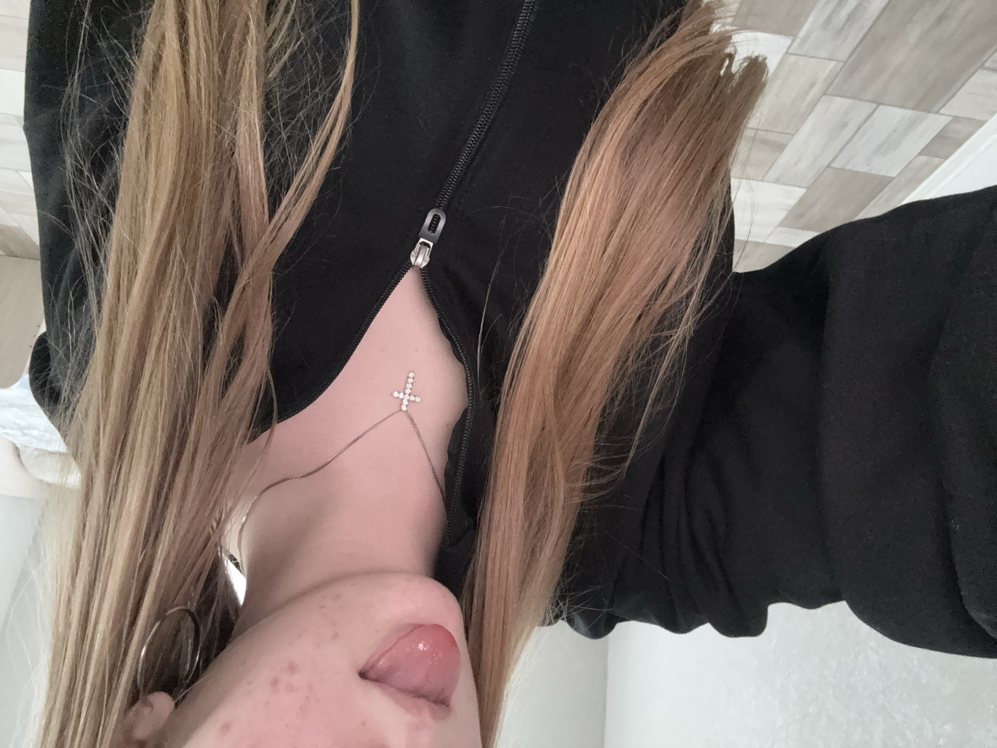 The image depicts a woman from an upside-down perspective. She has long, light brown hair and is wearing a black zip-up jacket. A silver necklace with a cross pendant is visible on her chest. Her tongue is slightly visible. The background shows a tiled wall and a white wall. The image doesn't provide information about location within a city. Therefore, it's not possible to confirm the location as Zhigalki, Russia.