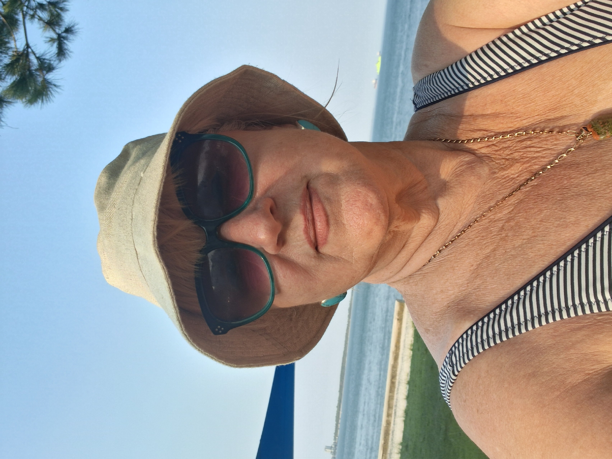 A woman in her 50s or 60s, with blonde hair, is taking a selfie outdoors. She is wearing a beige bucket hat, large sunglasses with green rims, and a striped swimsuit. She is also wearing a gold chain necklace with a pendant. The background shows a body of water, likely the Atlantic Ocean, with ships in the distance. A green grassy area and a concrete barrier are visible in the foreground. The sky is clear and blue. The woman's location is near the coast of Setúbal, Portugal.