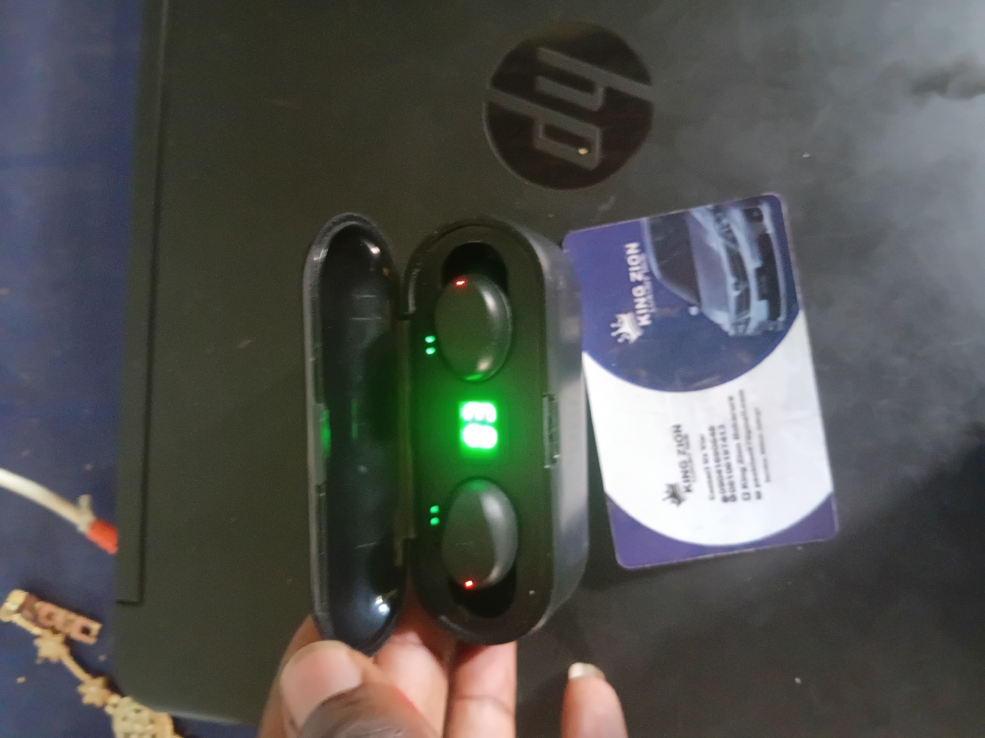 A dark-skinned hand holds an open black wireless earbud charging case, displaying green digital numerals "83" and two green indicator lights. A red indicator light is illuminated on the left earbud within the case. The case and a partially visible white and purple "KING ZION" business card rest on the closed lid of a black HP laptop, identifiable by its logo on the lower right. The business card details include contact information ("09041699646", "08106197413") and "King Zion Makurdi", along with an image of a silver sedan. In the upper left background, metallic debris and a white cable with a red tip are visible on a dark blue surface. This scene is located in the city of Jalingo, Nigeria.