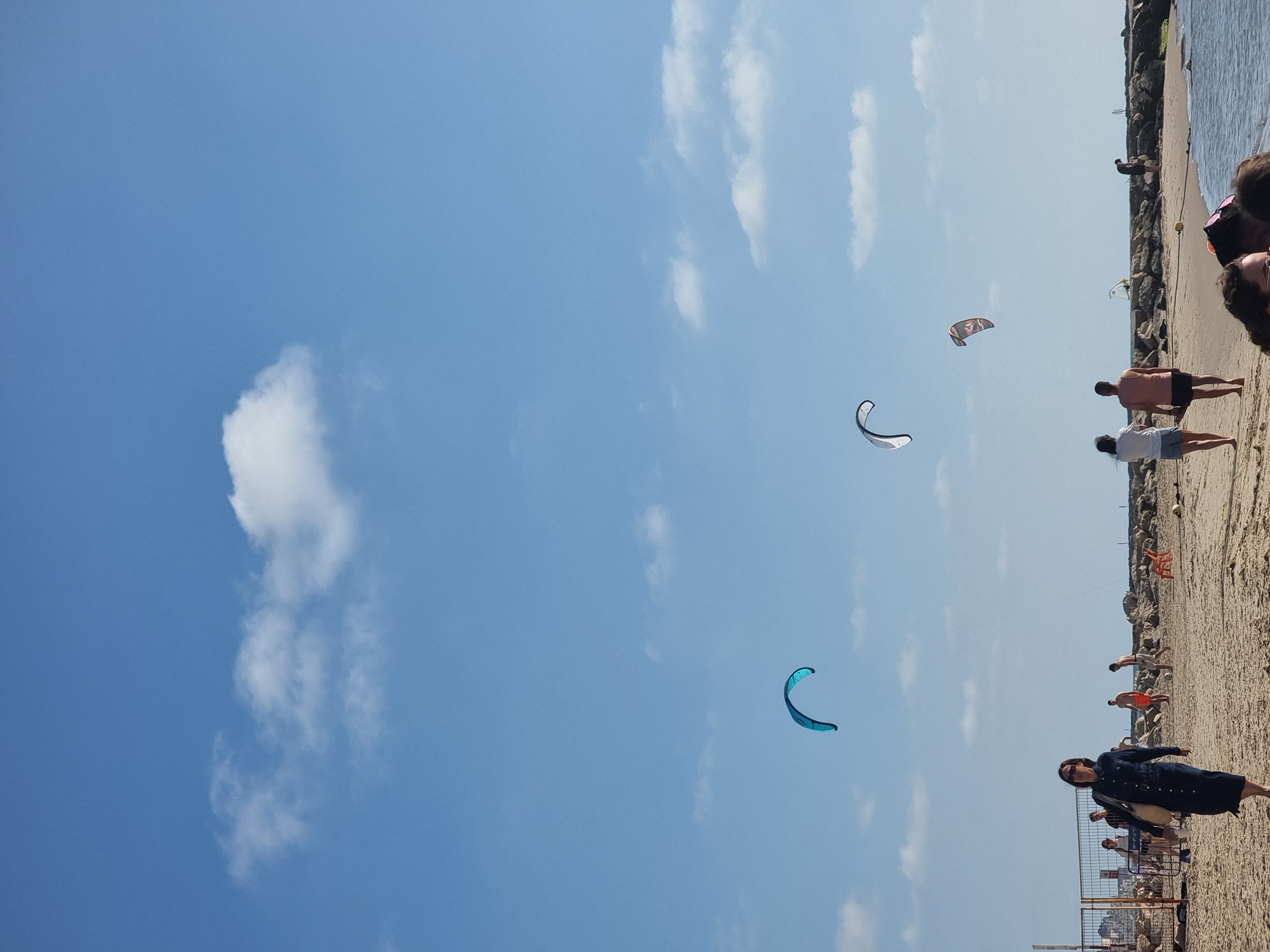 This image captures a vibrant, sunny day at a beach in Tel Aviv, Israel. The scene is dominated by a clear, bright blue sky with scattered white clouds, which fills the majority of the frame. On the right side, a sandy beach extends into the distance, meeting calm, blue ocean water. A rocky breakwater or jetty runs along the edge where the sand meets the sea on the far right.
Several individuals are present on the beach. In the immediate foreground on the right, a woman wearing a dark dress and sunglasses stands on the sand, looking towards the open sky. Further along the beach, two more people are standing, observing the scene; one wears a light-colored top and shorts, and the other a darker top and shorts. Distant figures can be seen further down the beach and near the water's edge, suggesting a lively environment.
The primary activity suggested by the image is kitesurfing or kiteboarding, indicated by three large kites prominently featured against the blue sky: one distinct turquoise kite, one white kite with black accents, and another red/orange and black/grey kite. A yellow buoy floats in the water near the shore. A metal fence or barrier is partially visible in the lower right, separating the immediate foreground beach area from a section behind it.
The time of day appears to be midday or early afternoon, characterized by strong, natural light and clear weather, perfect for outdoor and beach activities. The overall mood is one of leisure and active recreation. No discernible text is visible in the image.