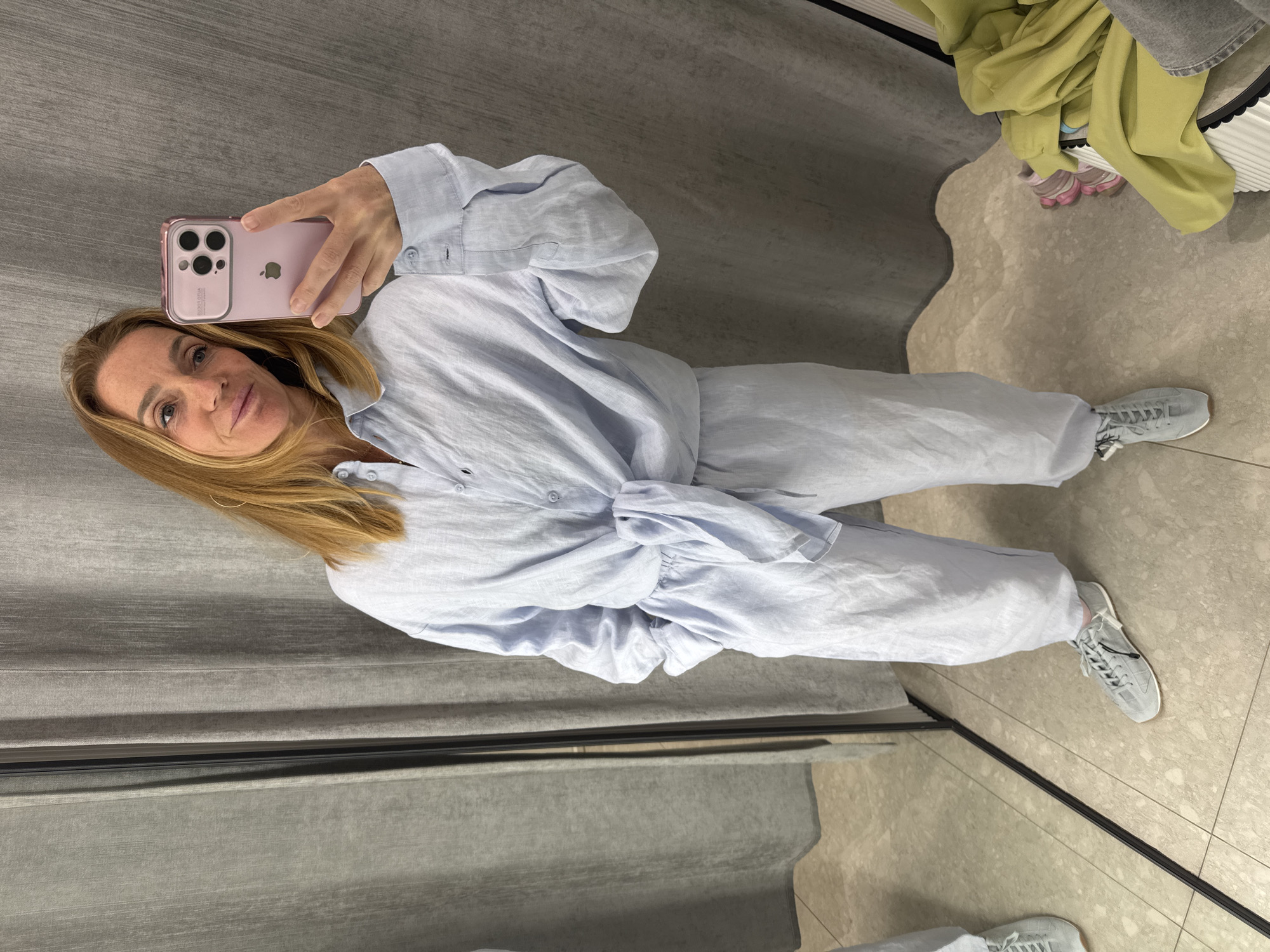 A woman is seen taking a mirror selfie in what appears to be a fitting room, likely in Saint Petersburg, Russia. She has long blonde hair and is looking directly into her pink iPhone, held up to capture her reflection, with a soft, pleasant smile. The phone's transparent case shows the "PRO" marking next to its camera lenses.
She is dressed in a light blue, two-piece linen outfit, featuring a long-sleeved button-up shirt tied at the waist, and matching wide-leg pants. Her feet are clad in grey sneakers with white soles. The setting is characterized by grey fabric curtains on the left and behind her, typical of a retail changing area. The floor is a light-colored tile with visible grout lines, overlaid with a wavy-edged, lighter-toned rug in the foreground. In the upper right background, a lime green garment and other clothing items are visible, possibly placed on a bench or shelf. Her reflection is visible in the mirror, including parts of her outfit and the room's interior.