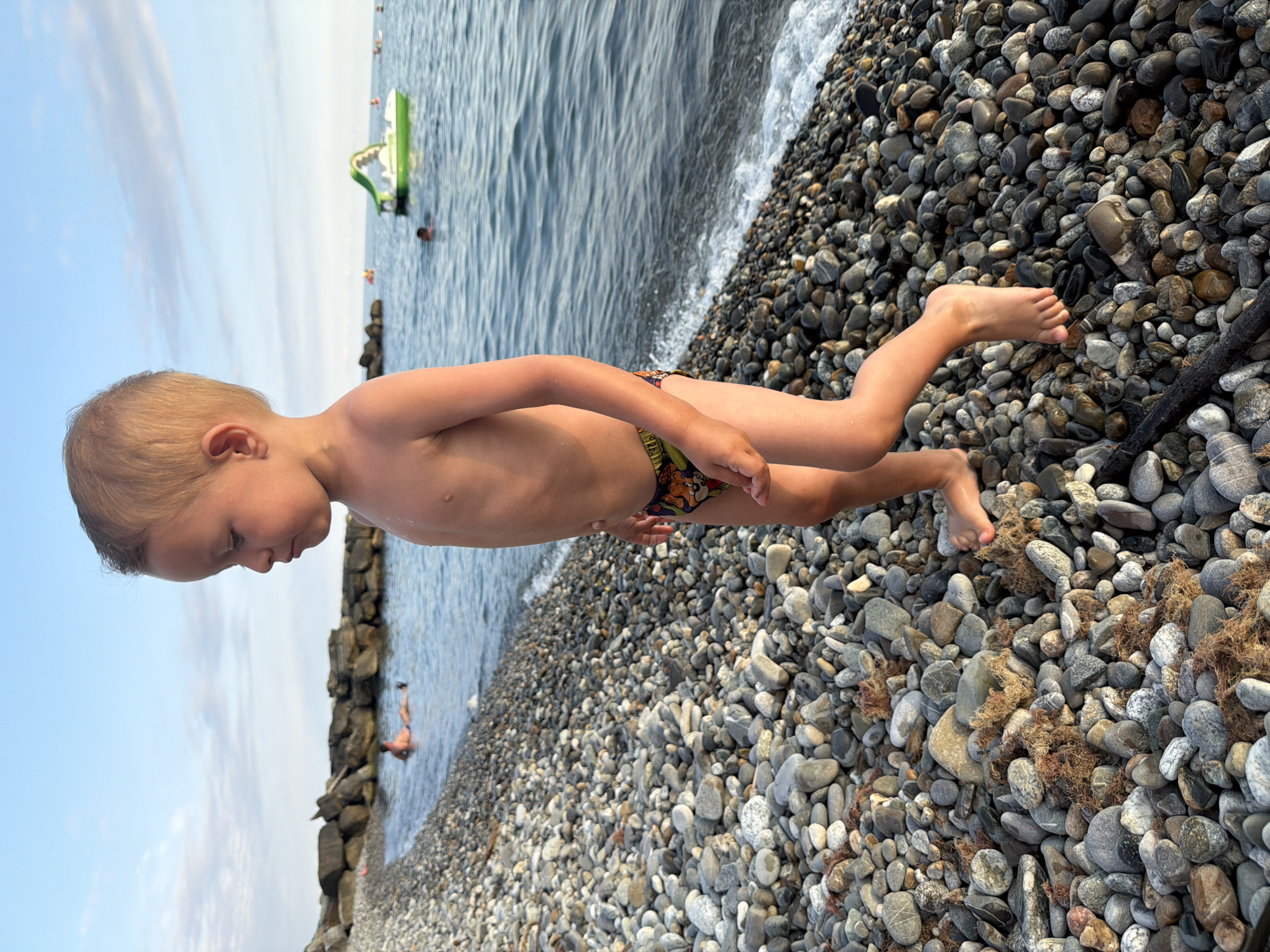 A young child walks on a pebble beach. The child is a toddler with blond hair, wearing patterned swimming trunks. The child is looking down and to the left, with their right leg extended forward. The beach is covered in smooth, grey and brown pebbles. The sea is calm with small waves breaking on the shore. In the background, there are large rocks forming a breakwater. Further out at sea, there are people swimming and a green pedal boat. The sky is clear with a few wispy clouds. The scene is likely a beach resort in Sochi, Russia, given the presence of a pedal boat and the overall setting.