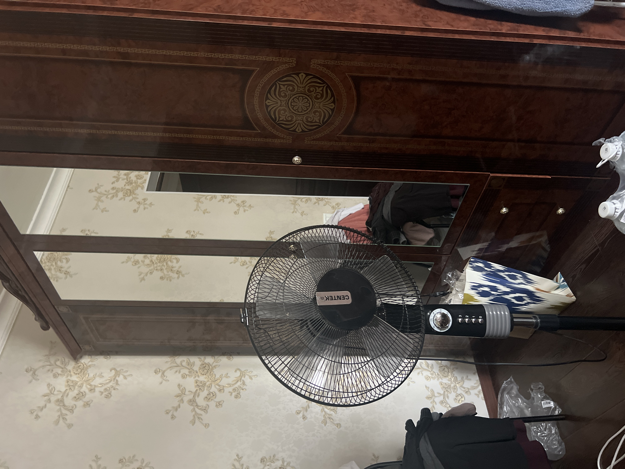 An indoor scene in Lysogorskaya, Russia, features a tall, dark wood wardrobe with mirrored doors and decorative inlays. To the left of the wardrobe stands a black oscillating floor fan, with its blades partially obscured by the cage. The fan's base is black, and a control panel with buttons and a dial is visible on its stem. Behind the fan, the wall is covered in wallpaper with a subtle floral pattern in a light color. On the floor, near the fan, are some plastic bags and a patterned shopping bag. Several items of clothing are piled on the floor to the left of the fan. The floor itself is made of dark wood planks.