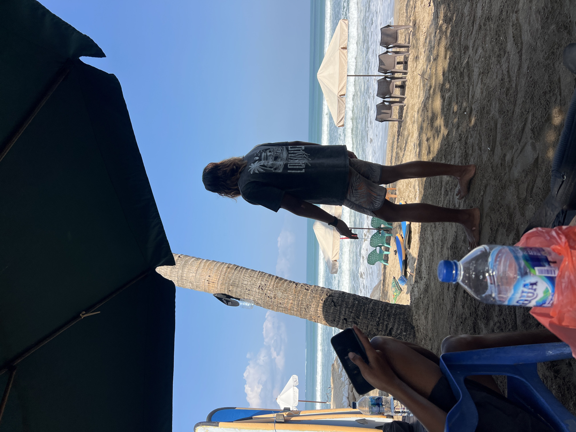 In Kuta, Indonesia, a man with long brown hair stands on a sandy beach, facing the ocean. He is wearing a dark t-shirt with a graphic design on the back and patterned shorts. The ocean, with gentle waves, stretches to the horizon under a clear blue sky. In the foreground, a large dark umbrella provides shade. To the left, a person in a blue chair holds a smartphone. Several beach chairs and umbrellas are set up on the sand. Surfboards are propped up near the left edge of the image. A bottle of Aqua water is visible in the foreground. A palm tree trunk is prominently featured near the center of the frame. The scene appears to be a relaxed day at the beach.