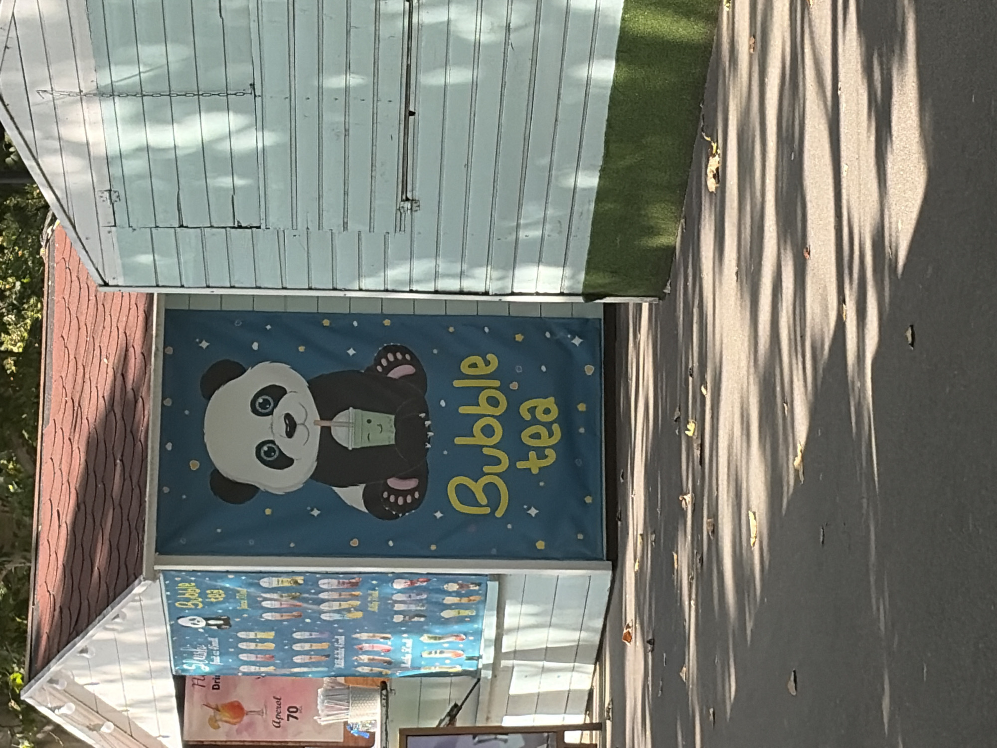 This is an outdoor, eye-level, medium shot of two small food kiosks. The kiosk on the left has a red shingled roof and white siding. A banner on the front features an illustration of a panda drinking bubble tea. Text on the banner reads "Bubble tea". Another banner to the left of it shows a cocktail and lists "Aperol 70". The kiosk on the right is painted a light blue and has wooden siding. A green strip of what appears to be artificial turf runs along the base of this kiosk. The ground in front of the kiosks is paved and scattered with fallen leaves. The scene appears to be well-lit by sunlight, casting shadows from trees. There are no people visible in the frame. The location is identified as Chișinău, Moldova.