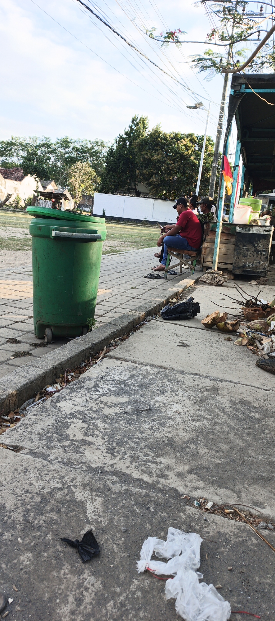 In Kasiyan Timur, Indonesia, a scene unfolds on a paved outdoor area. A large green trash can with a lid rests on wheels to the left. In the background, a wooden kiosk stands, displaying various items. Two individuals are seated on chairs in front of the kiosk, one looking at a mobile phone. The ground is a mixture of paving stones and concrete, with debris such as coconut husks, dry leaves, and discarded plastic bags scattered about. Overhead, power lines crisscross against a clear sky.