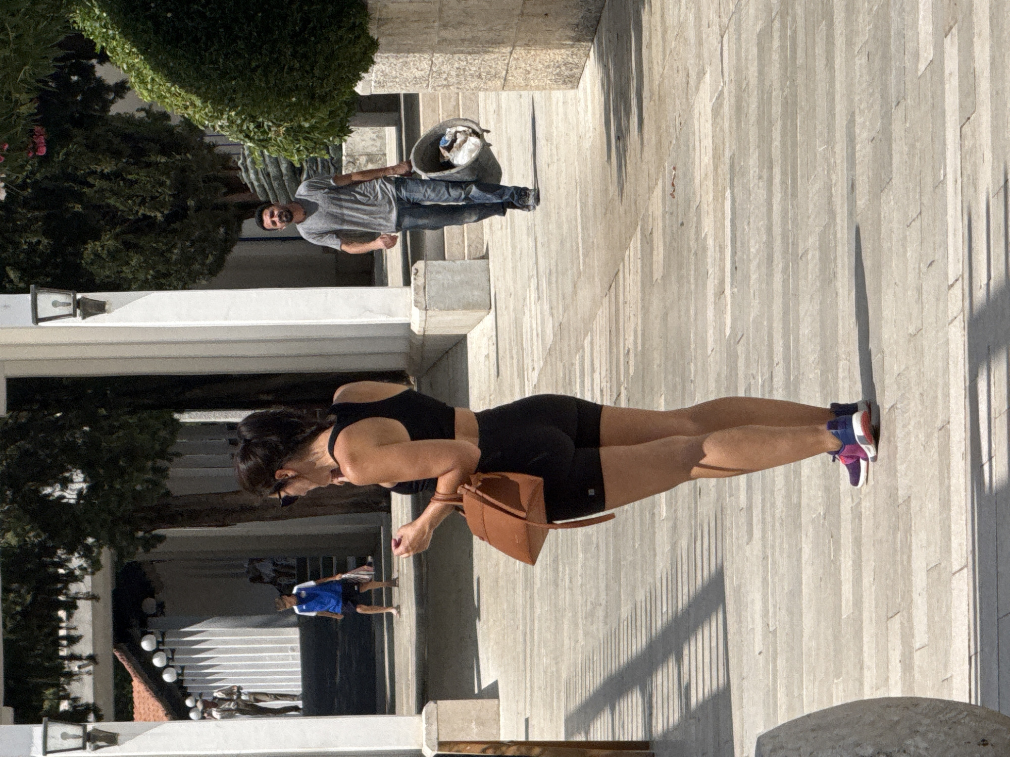 In Boreti, Montenegro, a woman in black athletic wear, including a sports bra and shorts, stands facing away from the camera, holding a tan handbag. She is wearing sunglasses and multi-colored sneakers. In the background, a man walks towards the viewer, carrying a large gray bucket filled with debris. He is wearing a gray t-shirt and blue jeans. Further in the background, a young boy in a blue and white soccer uniform walks with a plastic bag. The scene takes place outdoors on a paved walkway with architectural elements and trees visible.