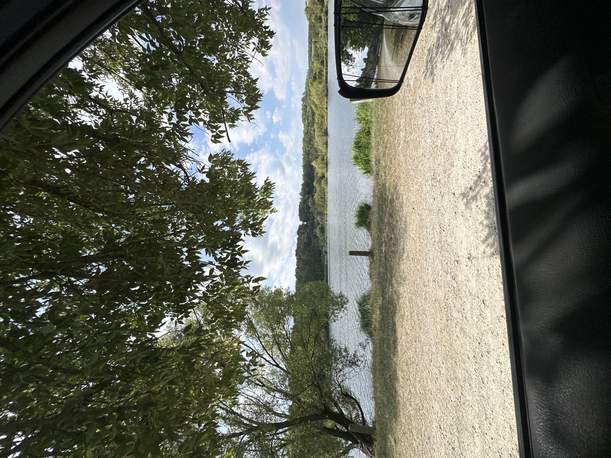The image is a view from inside a vehicle, looking out at a serene lakeside scene. Large green leaves from a tree in the foreground partially obscure the view of a calm body of water, which stretches towards a tree-lined horizon. The sky above is a bright blue, dotted with fluffy white clouds. On the right side of the frame, a side-view mirror of the vehicle reflects the surrounding environment. The ground in front of the water appears to be a sandy or gravelly area. The image is likely taken in the vicinity of Makiv, Ukraine, given the natural landscape and the presence of water.