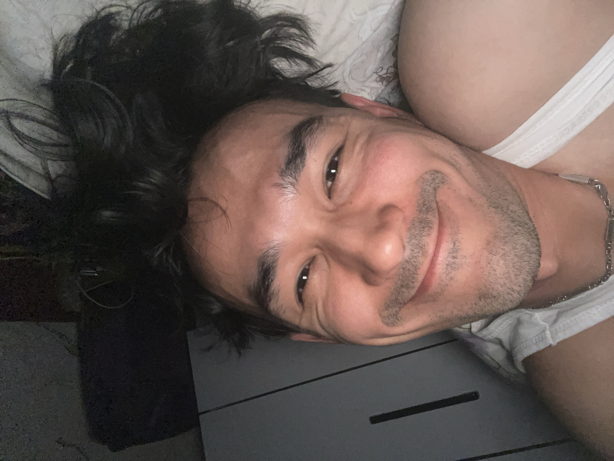 A close-up, low-angle, dimly lit, over-the-shoulder selfie shows a person lying on a bed. The person is partially visible, with their head turned towards the camera. They have dark hair, a fair complexion, and are wearing a white tank top. Their facial expression is a gentle smile. The background appears to be a bedroom with a dark dresser and a white blanket. The image is oriented sideways. There is no discernible information to suggest the location is Daşoguz, Turkmenistan.