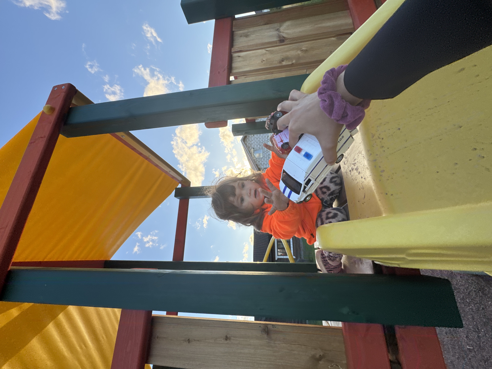 A young child with brown hair, wearing an orange hooded jacket and patterned leggings, sits on a yellow plastic slide on a playground. The child's hands are outstretched as if reaching for or playing with toys. A person's hand, wearing a purple scrunchie on their wrist, is visible in the foreground, holding a white toy police car with red and blue lights. Another small toy car is also visible. The playground structure has wooden and red elements, with a yellow fabric canopy overhead. The background shows a clear blue sky with scattered clouds and a building with a pitched roof, suggesting a residential area. The overall scene depicts a moment of play outdoors.