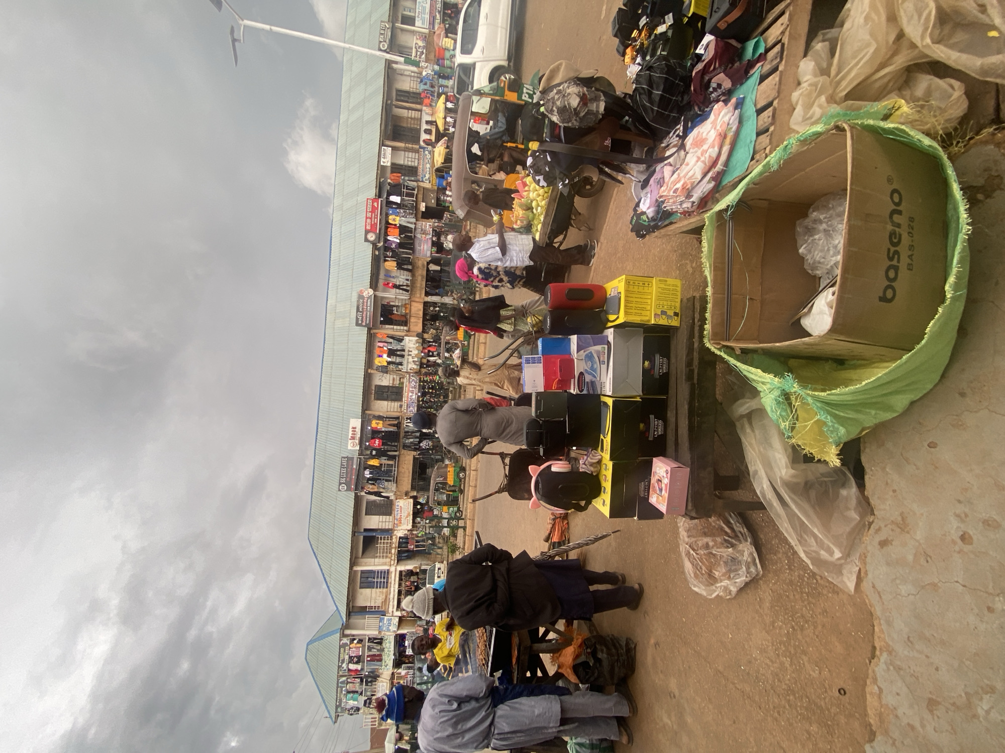 The image depicts an outdoor market scene in Bukuru, Nigeria. In the foreground, a collection of electronic items, including speakers and boxed devices, are displayed on a table and the ground. To the left, two men in traditional attire are engaged in a transaction at a food stall, with a grill of meat visible. Behind them, a vendor wearing a yellow shirt attends to customers. A third man in a gray tunic stands to the right, facing away from the camera. Further into the scene, several people are browsing merchandise displayed on racks outside storefronts. The buildings appear to be shops with awnings and signs, one reading "SK LUXE LANE." Various vehicles, including a trishaw and a white pickup truck, are present on the street. The sky is overcast with gray clouds. The ground is a mix of dirt and pavement. Several plastic bags and a large green sack containing items are also visible.