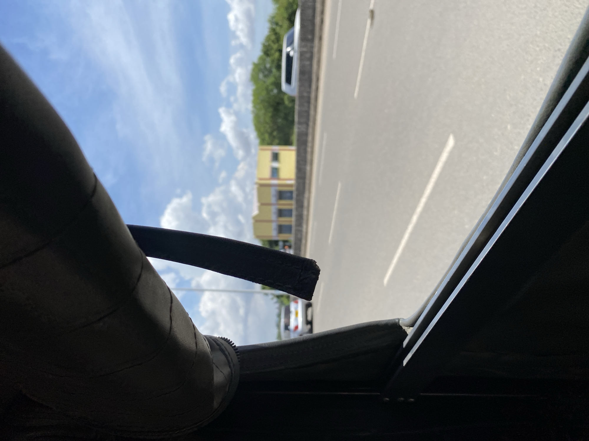 The image shows a view from inside a vehicle, looking out towards a road in Dar es-Salaam, Tanzania. A fragment of the vehicle's interior, including a roof support and trim, is visible in the foreground. The road appears to be a highway with multiple lanes. Other vehicles can be seen on the road, including a white car in the distance and another partially obscured vehicle further ahead. A large yellow building is visible alongside the road, possibly a commercial or residential structure. The sky is partly cloudy with blue patches.