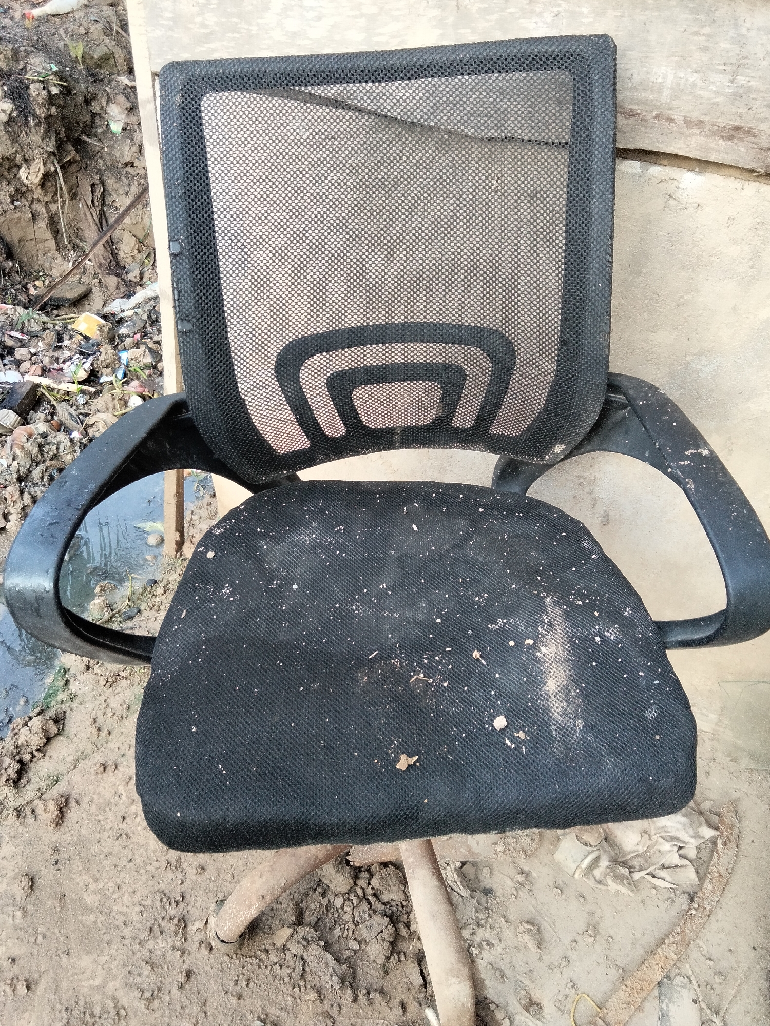 The image depicts a black mesh office chair that is in a dilapidated state. The seat of the chair is covered in a significant amount of dirt and debris, and the mesh on the backrest appears torn and damaged. The chair is situated outdoors on a muddy surface, with visible trash and murky water in the background, suggesting an unkempt or neglected environment. The overall impression is one of abandonment and decay, potentially in an outdoor area within Buleleng Regency, Bali, Indonesia.