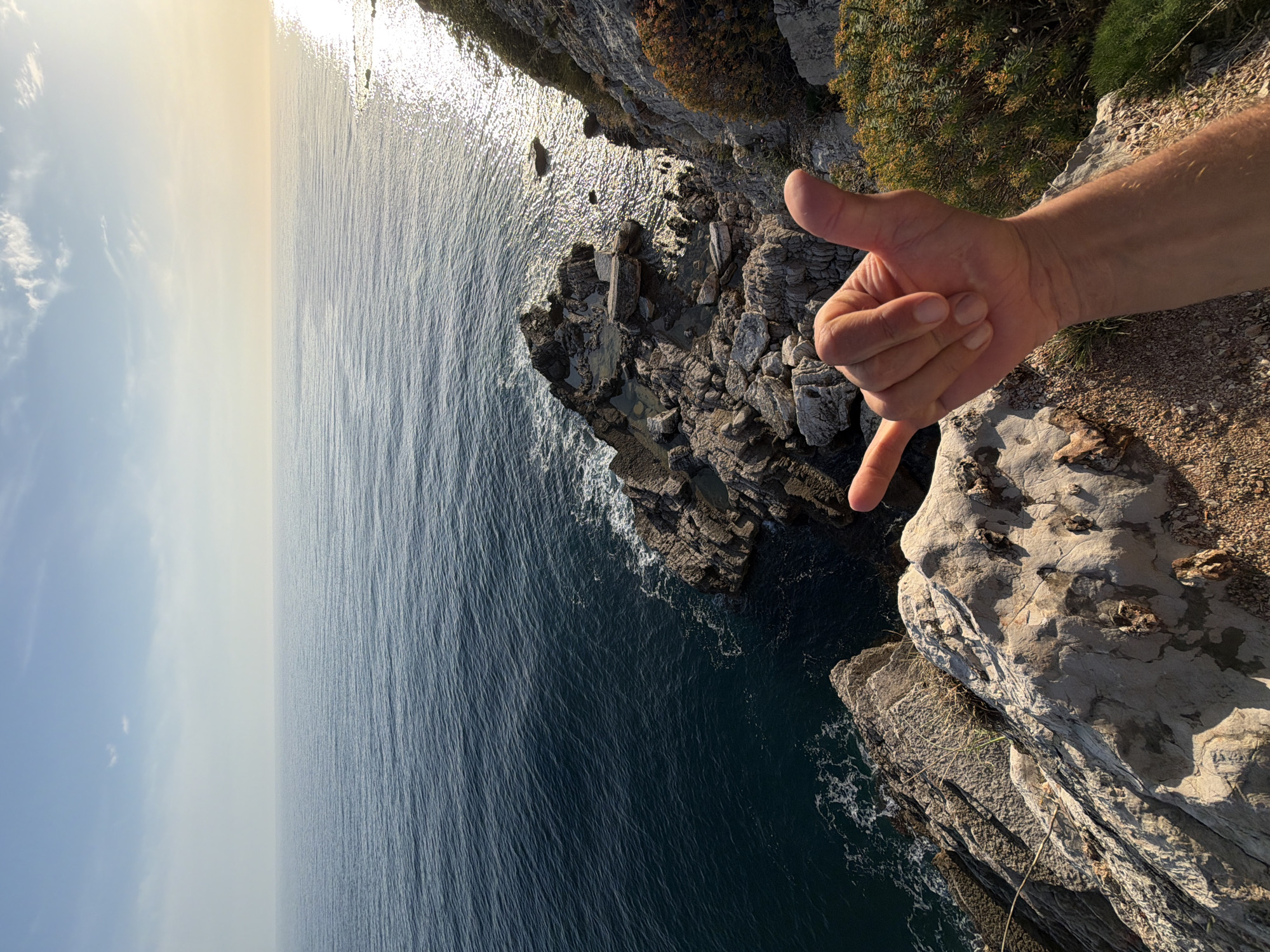 The image shows a coastal scene in Đurovići, Montenegro, viewed from a cliff edge. The Adriatic Sea stretches to the horizon, reflecting sunlight. The rocky cliff face drops steeply into the sea. A person's arm and hand are extended into the frame, gesturing with a "hang loose" sign. Vegetation is visible on the cliff edge.