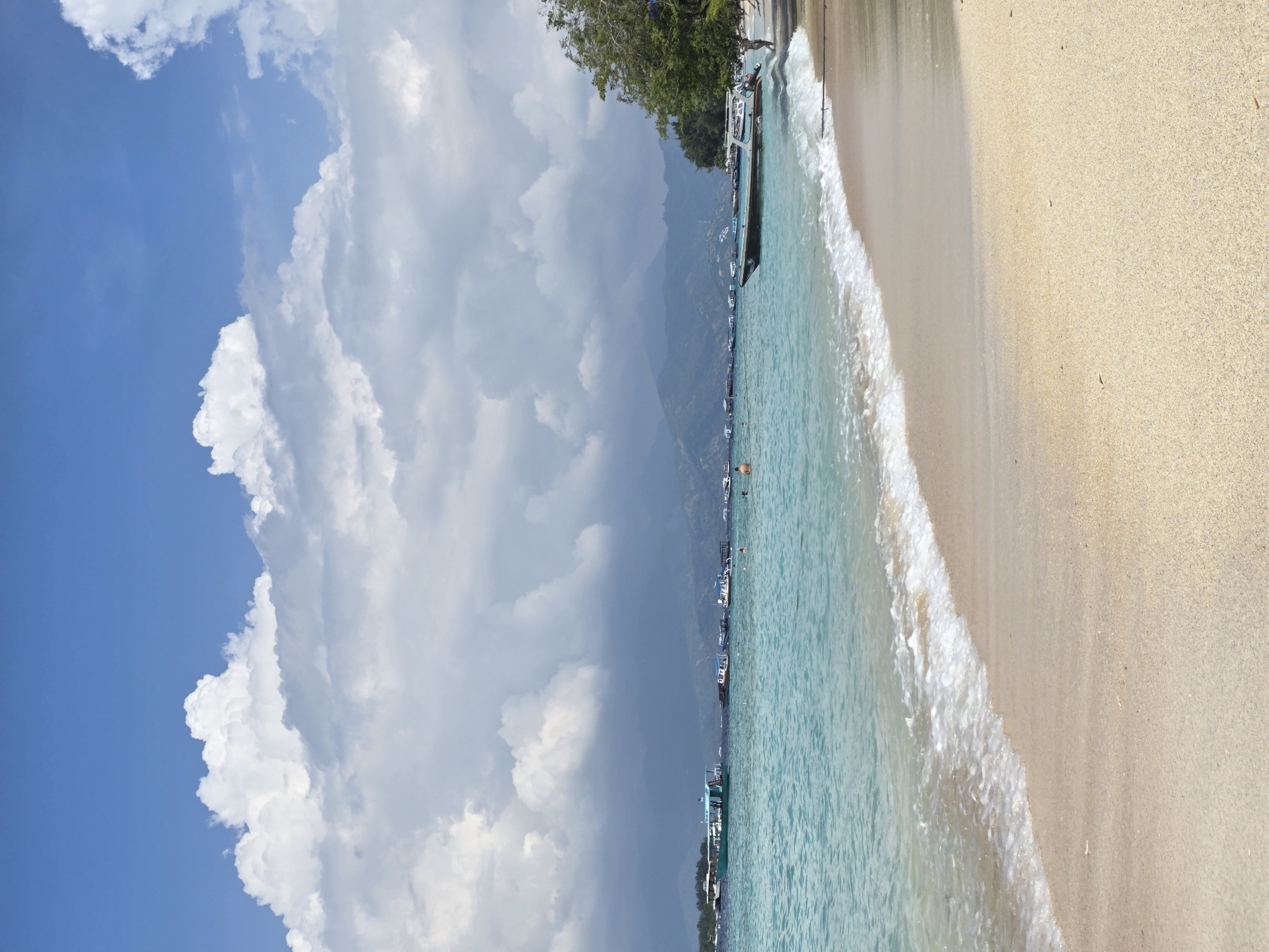 The image captures a beach scene. The location is a beach in Unknown, Indonesia. The photo is oriented vertically.
In the foreground, there is a sandy beach, with a clear separation between the sand and the water. Waves are visible as they break onto the shore.
The water is clear and turquoise in color. A line of boats is anchored near the shore. Further out, individuals can be seen swimming in the water.
In the background, there is a mountain range, partially obscured by clouds. The sky is blue with large, puffy white clouds.