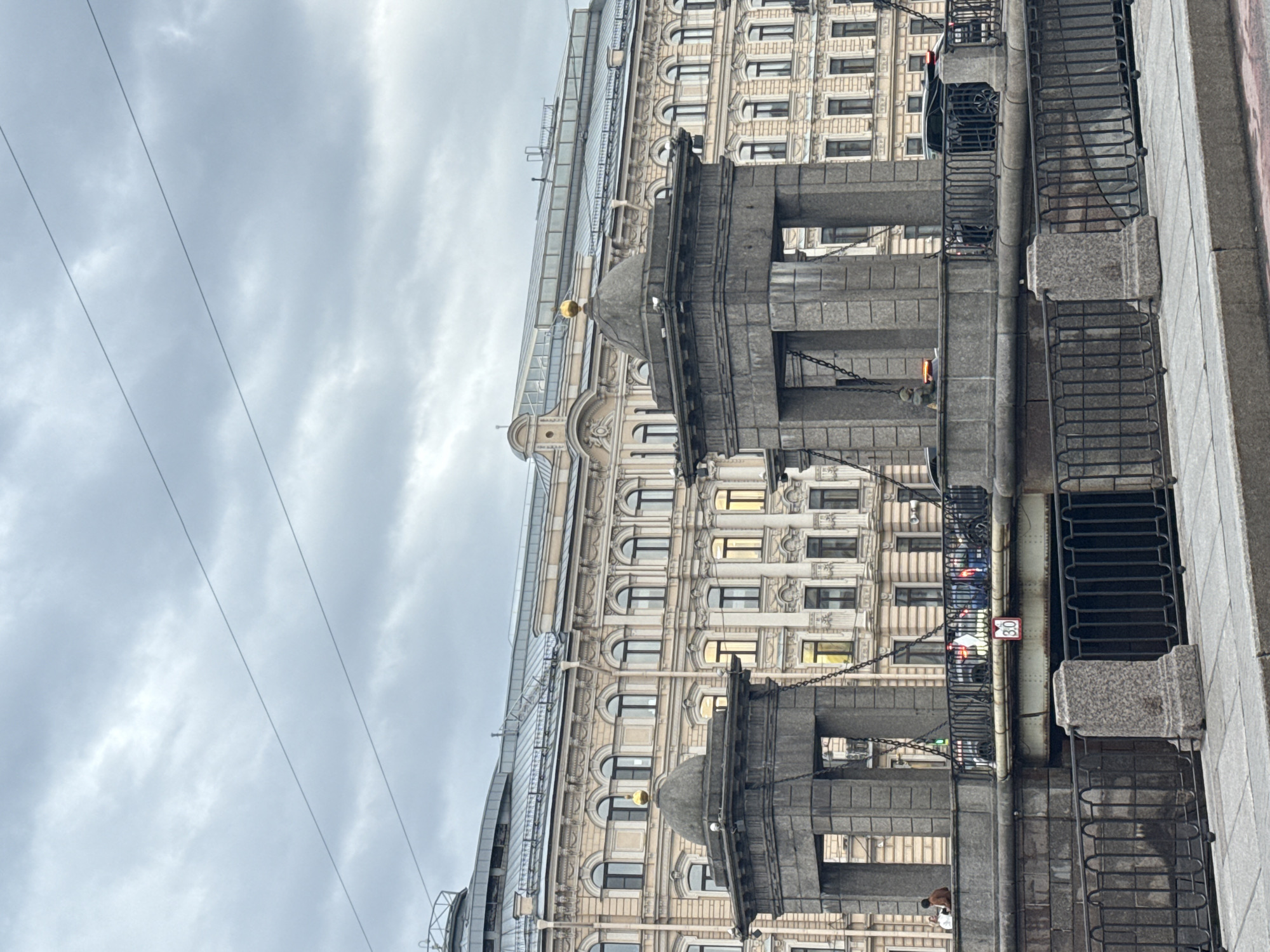 The image captures a street scene in Saint Petersburg, Russia, showcasing architectural elements of the city. The foreground features a bridge structure with dark metal railings and granite-like stone supports. Behind the bridge, a grand, multi-story building rises, exhibiting an ornate facade with numerous arched windows and decorative moldings. Some windows are illuminated from within, suggesting interior lighting. Above, a cloudy sky casts a muted light over the scene. A few power lines run across the upper left portion of the frame. In the lower part of the image, under the bridge structure, a person is visible.