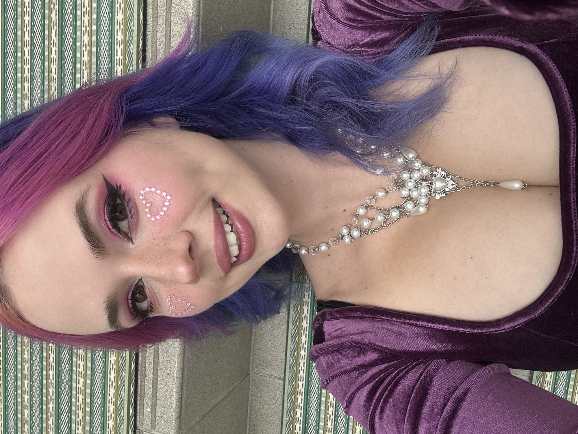 A close-up, tilted portrait features a young individual smiling towards the camera, showcasing a cheerful expression with slightly parted lips revealing teeth. The person has vibrant, two-toned hair, transitioning from pinkish-purple at the front to blue-purple towards the back, styled with soft waves. Their face is adorned with intricate makeup, including pink eyeshadow, sharp winged eyeliner, and distinctive heart-shaped embellishments made of small, pearl-like beads on their cheekbones. Subtle freckles are visible across their nose and cheeks.
The subject is wearing a deep purple, possibly velvet, garment with a low-cut neckline and long sleeves, suggesting a stylish outfit. A prominent multi-strand pearl and silver-toned necklace, featuring an ornate central pendant and a hanging pearl drop, accessorizes their décolletage.
The background consists of two distinct sections: on the left, a vertically striped pattern in shades of green, white, and brown, resembling a woven textile or decorative panel; on the right, a plain gray surface, possibly concrete or stone. The overall composition suggests a posed portrait, highlighting the subject's unique aesthetic. The event takes place in the City of Zagreb, Croatia. No text is visible in the image.