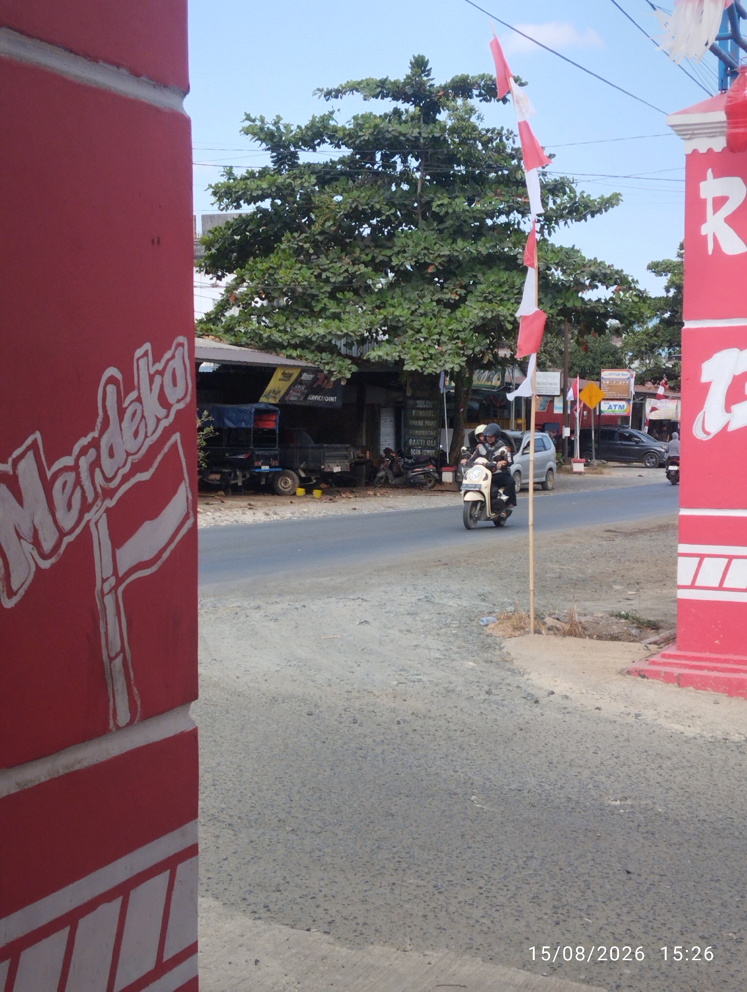 The image depicts a street scene in Tegal Rejo, Indonesia. In the foreground, a red pillar with white lettering that reads "Merdeka" and a stylized Indonesian flag is visible. Red and white Indonesian flags are also hanging from a pole to the right of the pillar, and attached to wires above the street. A tree with dense green foliage stands behind the flags. On the street, two people are riding a scooter, and further down the road, a pickup truck is parked under a canopy. Several other vehicles, including a silver car and a dark SUV, are also present on the road. The environment appears to be a commercial area, with buildings and signs visible in the background. The road surface is asphalt, and the sky is clear and blue. A timestamp in the lower right corner indicates the image was taken on 15/08/2026 at 15:26.
