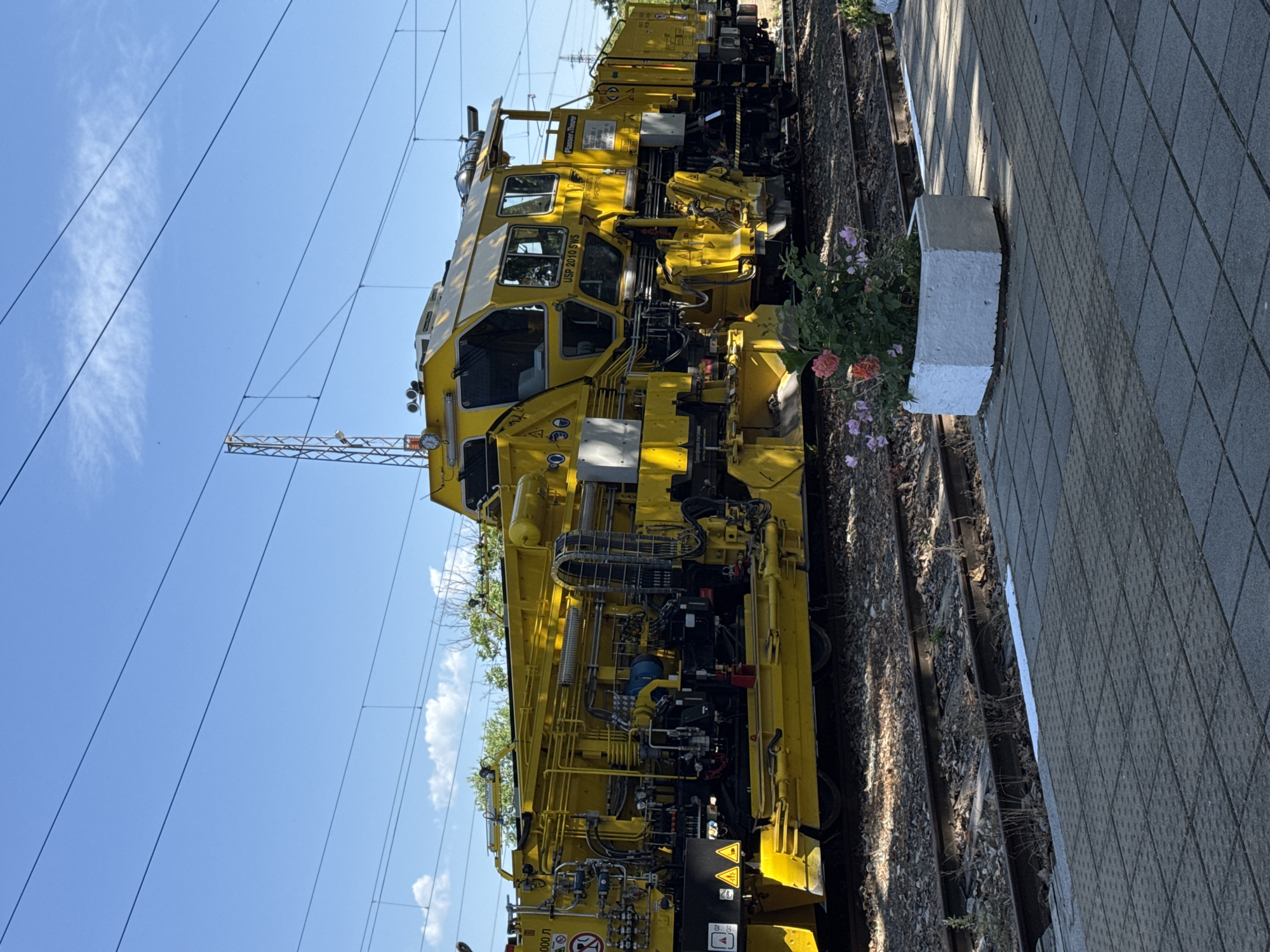 A yellow Plasser & Theurer track maintenance machine, specifically labeled as "USP 2010 SWS," is positioned on railway tracks adjacent to a paved platform. A white concrete planter box with flowers sits between the tracks and the platform edge. Overhead power lines are visible against a clear blue sky. The image is oriented sideways, showing the machine and platform in a vertical alignment. The city is Kaloyanovets, Bulgaria.