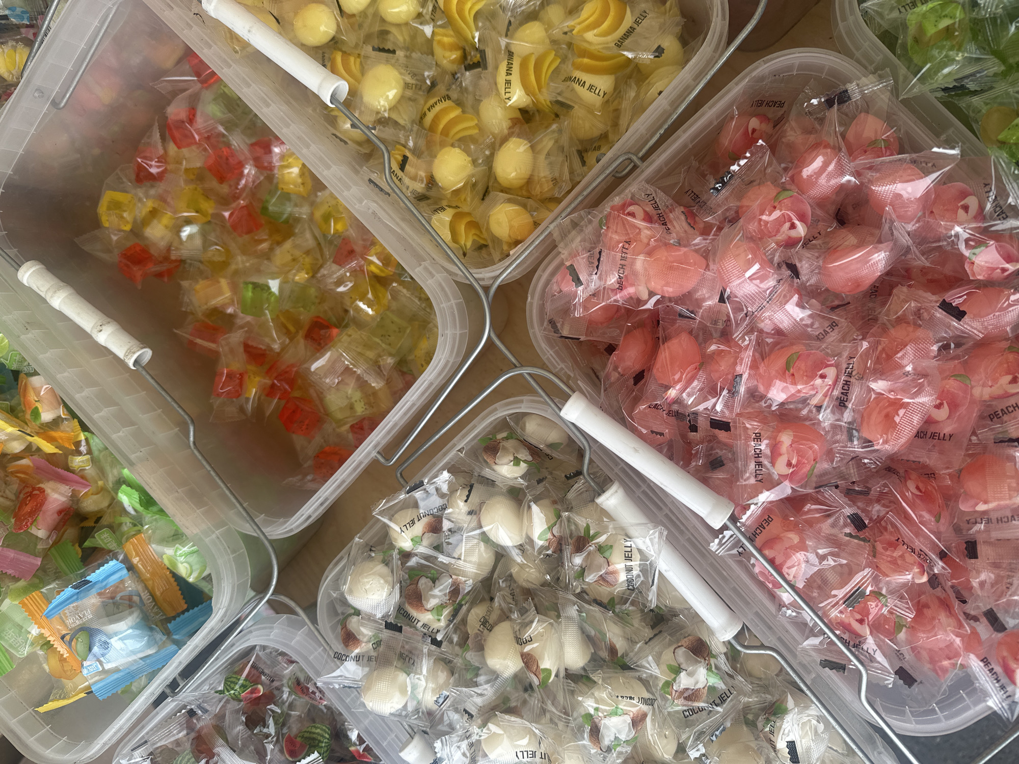 The image presents a high-angle shot of five transparent plastic containers arranged closely together, each filled with individually wrapped jelly candies. The candies come in various shapes and flavors, including "banana jelly," which appears as yellow banana and round shapes; "coconut jelly," shaped like white balls; "peach jelly," shaped like peaches and colored in light pink and red; and a mix of red, yellow, and green cube-shaped jelly candies. The fifth container contains a variety of smaller candies. The containers have white handles and are placed on a neutral-colored surface, which seems to be a table or counter. The scene is well-lit, and the focus is sharp, providing a clear view of the candy and packaging details. The context of the image is likely a candy store or market stall. There is no indication of the location being Chaikovsky, Russia in the picture.