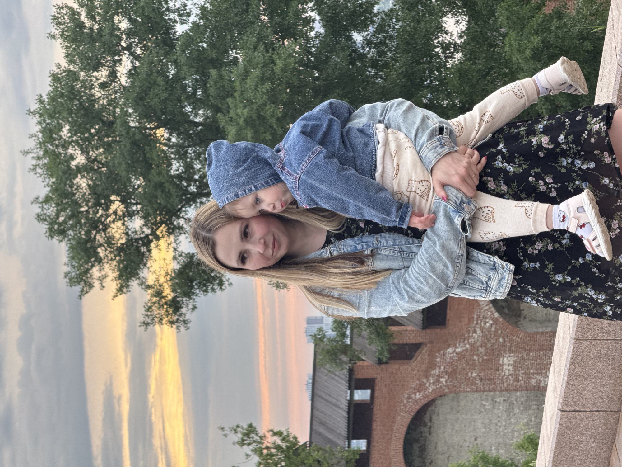 The image depicts a woman holding a young child in front of a sunset sky. The woman has light brown hair and is wearing a denim jacket and a black floral dress. The child is wearing a matching denim jacket and hat, with beige leggings featuring a cat pattern. The backdrop includes greenery, a segment of a brick building with an arched entrance, and a stone wall. The setting is identified as Nizhny Novgorod, Russia, based on the architecture of the brick building.