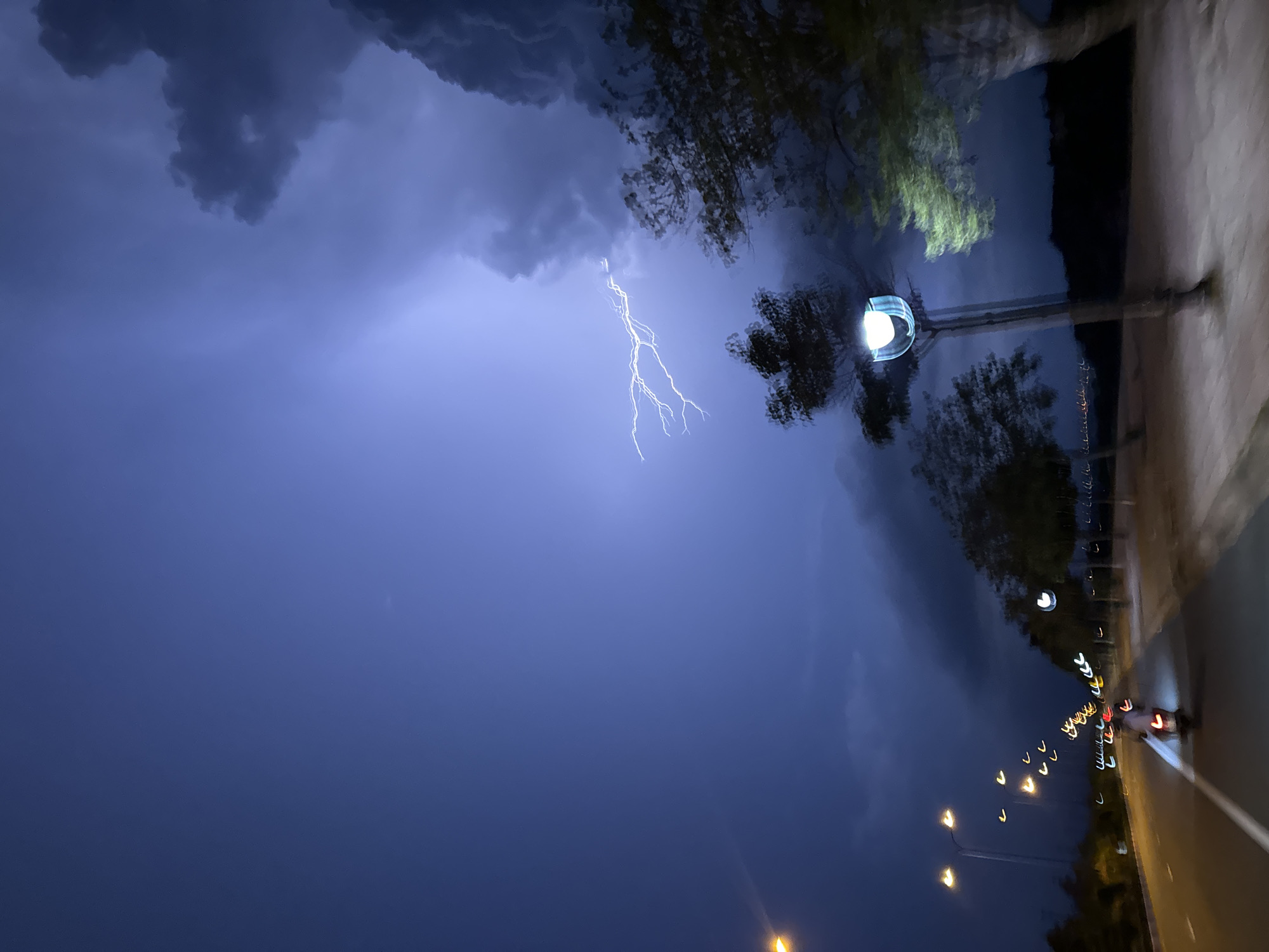 A photograph shows a lightning strike over the Phu Thuy Ward in Vietnam at night. The sky is filled with dark, stormy clouds, illuminated by a bright flash of lightning. The background features trees, and street lights lining a road. A street lamp is visible in the foreground.