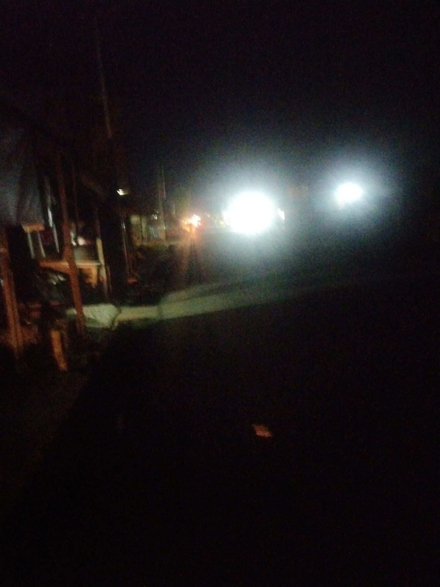 The image depicts a dimly lit street scene at night, possibly in Uyo, Nigeria. On the left, there's a structure with a makeshift appearance, constructed with wooden elements. A faint white covering or object is also visible within this structure. In the distance, two bright lights illuminate the scene, suggesting the presence of a vehicle. Additionally, there are a few other lights or indistinct objects in the distance that could be buildings or streetlights. The overall environment appears dark and the details are somewhat obscured by the low light conditions. The lower foreground of the image is heavily shadowed.