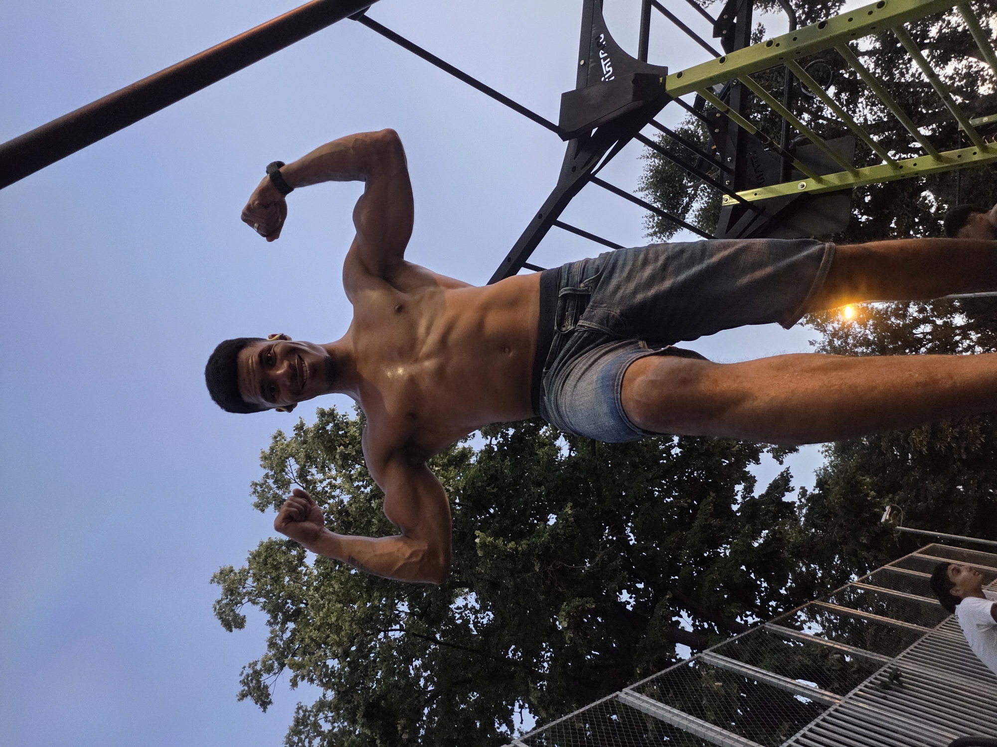 A low-angle shot shows a shirtless young man flexing his biceps outdoors, with a clear sky overhead. He is wearing denim shorts. Behind him, there are trees and a metal climbing structure. To the left, a chain-link fence encloses an area. The man appears to be in a park or outdoor gym. The setting resembles an urban park, possibly in Varna, Bulgaria, given the presence of urban infrastructure like the fence and climbing equipment.
