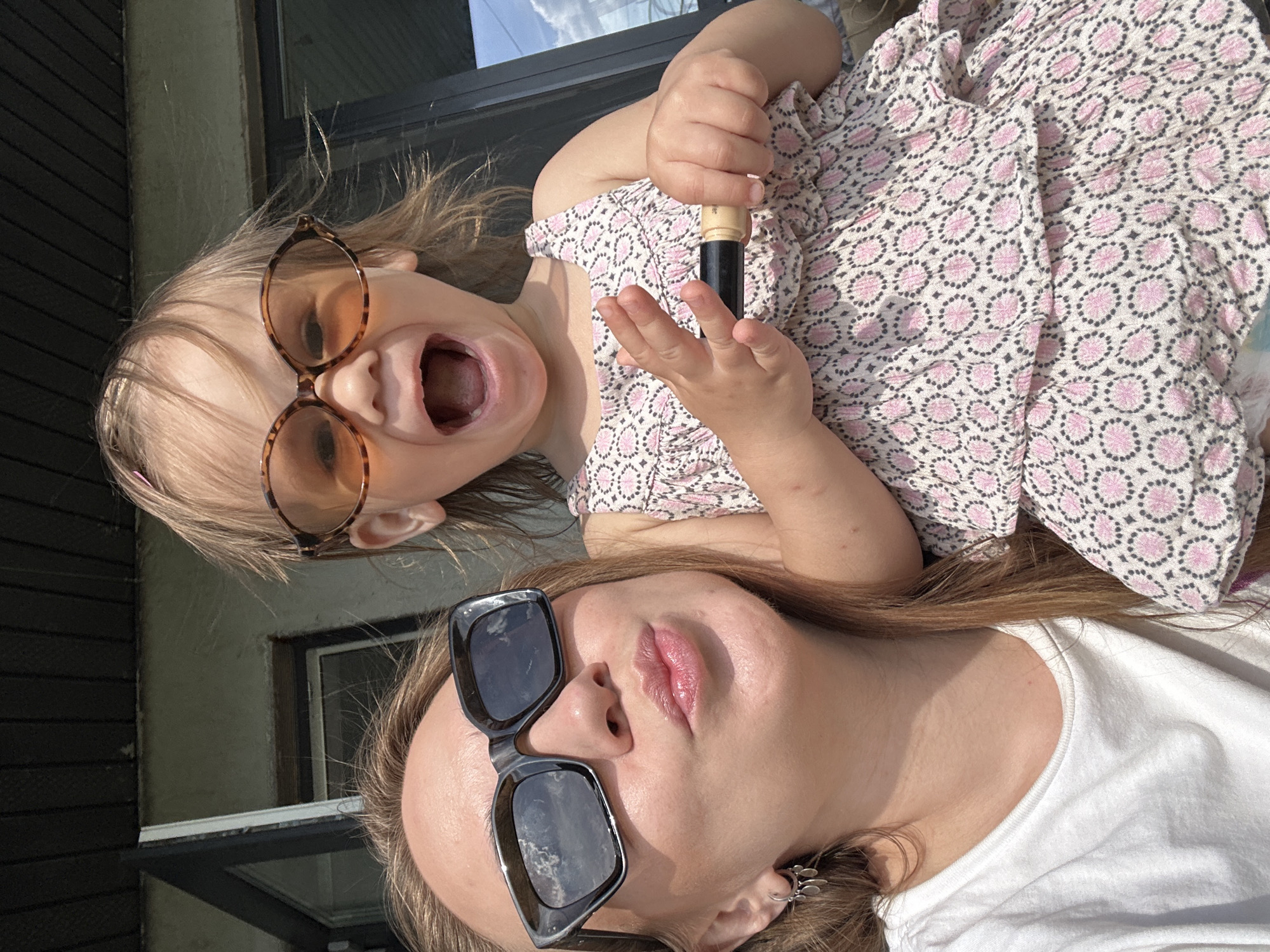 A medium shot captures two figures against an outdoor backdrop. An adult, wearing oversized square black sunglasses and a white t-shirt, is positioned in the foreground. Above them, a child with fair hair wears round, spotted sunglasses, a dress with a pink and black patterned bodice, and holds a dark-colored object with a tan handle. The child has their mouth open in a shout. In the background, architectural elements are visible, suggesting a building or structure, though the specific location in Kharnavy, Russia is not discernible. The image composition suggests a selfie or a photo taken at close range.