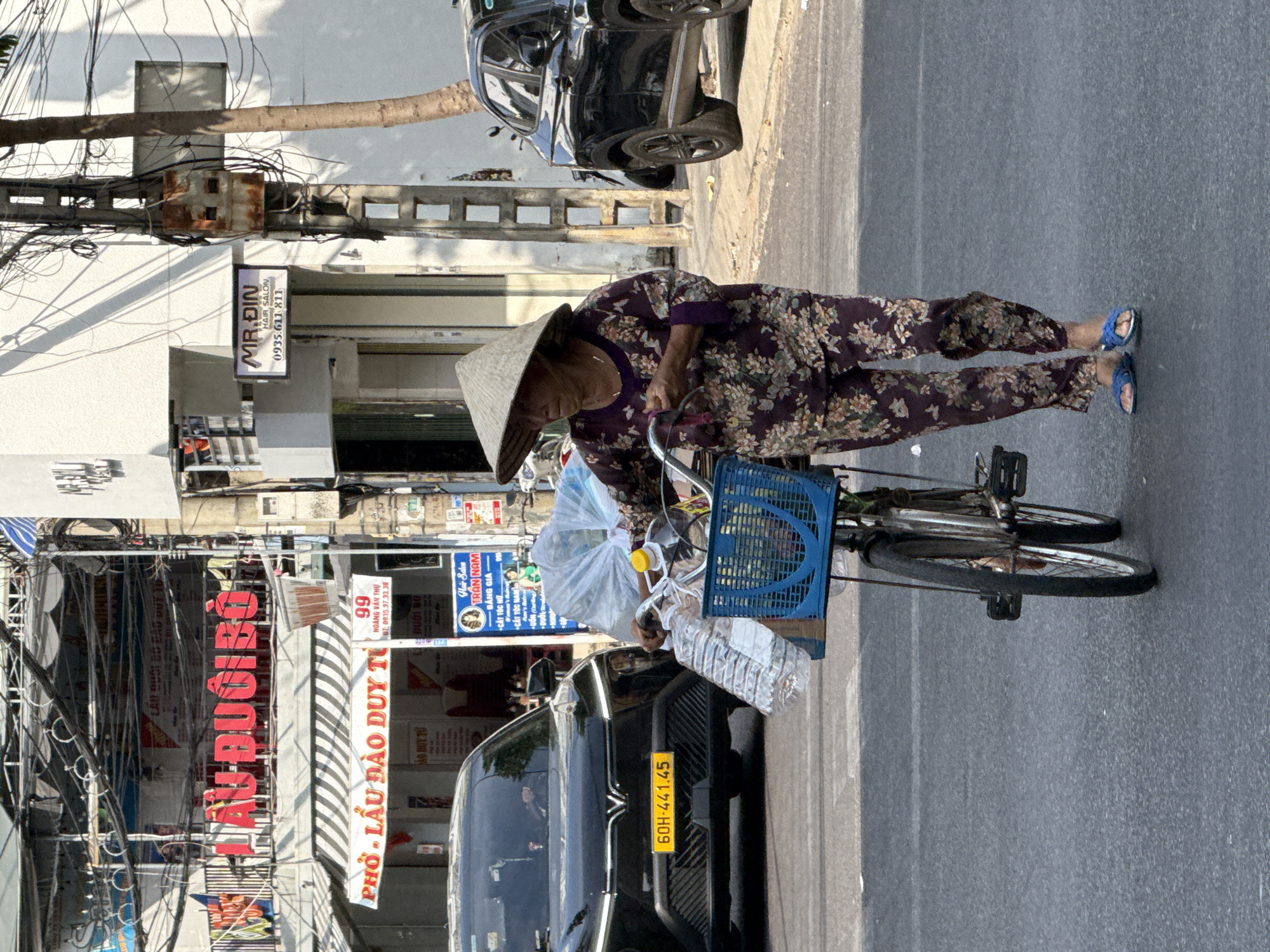 An elderly woman, wearing a traditional Vietnamese conical hat (nón lá) and a purple floral-patterned outfit, stands next to her bicycle on a sunlit street in Nha Trang, Vietnam. She appears to be tending to items on her bicycle, which include a blue plastic basket filled with plastic bags and water bottles. She wears blue sandals on her feet.
The setting is an urban street featuring buildings, utility poles, and an intricate network of overhead wires. A black car with a Vietnamese license plate "60H-441.45" is parked nearby. The bright sunlight casts distinct shadows, indicating midday or early afternoon on a clear day.
Several business signs are visible:
- "MR.DIN HAIR SALON" with the phone number "0935.611 811".
- "PHỞ - LẦU ĐÀO DUY TỪ 99" with the phone number "ĐT: 0935.37.33.38", indicating a restaurant serving pho and hotpot.
- A sign for "Hair Salon TRAN NAM" listing services: "BẢNG GIÁ" (price list), "CẤY TÓC NỮ" (women's hair transplant), "CẮT TÓC NAM" (men's haircut), and "UỐN" (perm).
- Partial text "DƯỚI BÓ" is also visible on a sign.