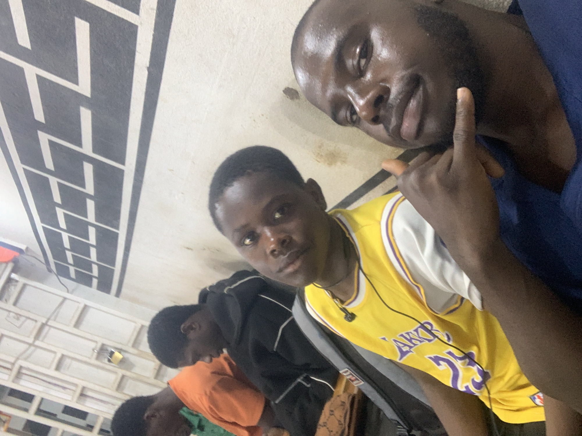 A selfie shows five young men indoors. The man in the foreground wears a yellow Lakers jersey with the number 24. Behind him, a man is dressed in a black hoodie. Another individual is wearing an orange shirt. The fifth man is wearing a blue shirt. The lighting is dim, and the setting appears to be a simple interior with a patterned accent strip on the wall. The location is Jos, Nigeria.