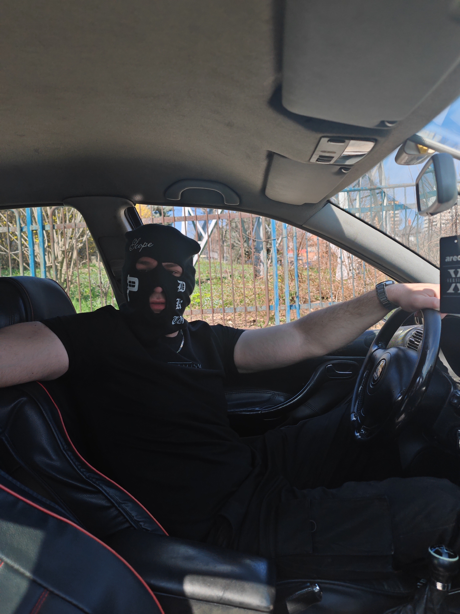 An individual is observed seated in the driver's seat of a passenger vehicle during daylight in Shumen, Bulgaria. The person is wearing a black balaclava with white text, "Scope" on the forehead and stylized "D.K.O.S." on the right side of the face. The individual is dressed in a plain black t-shirt, black cargo pants, a silver-colored neck chain, and a silver-colored wristwatch on the left wrist. The left arm rests on the vehicle's interior door panel, while the right hand loosely grips the black four-spoke steering wheel.
The vehicle's interior features a dark grey fabric headliner, an overhead console with lights, and sun visors in the raised position. The front passenger seat, partially visible, is upholstered in black faux leather with red contrast stitching. An "areon" brand air freshener hangs from the rearview mirror. A manual gear shifter is visible to the right of the driver.
Visible through the vehicle's windows is an exterior scene featuring a metallic fence composed of vertical blue bars and horizontal yellow-brown bars. Beyond the fence, leafless trees and some sparse green vegetation are present under a clear blue sky, indicating an outdoor, possibly urban or suburban, environment.