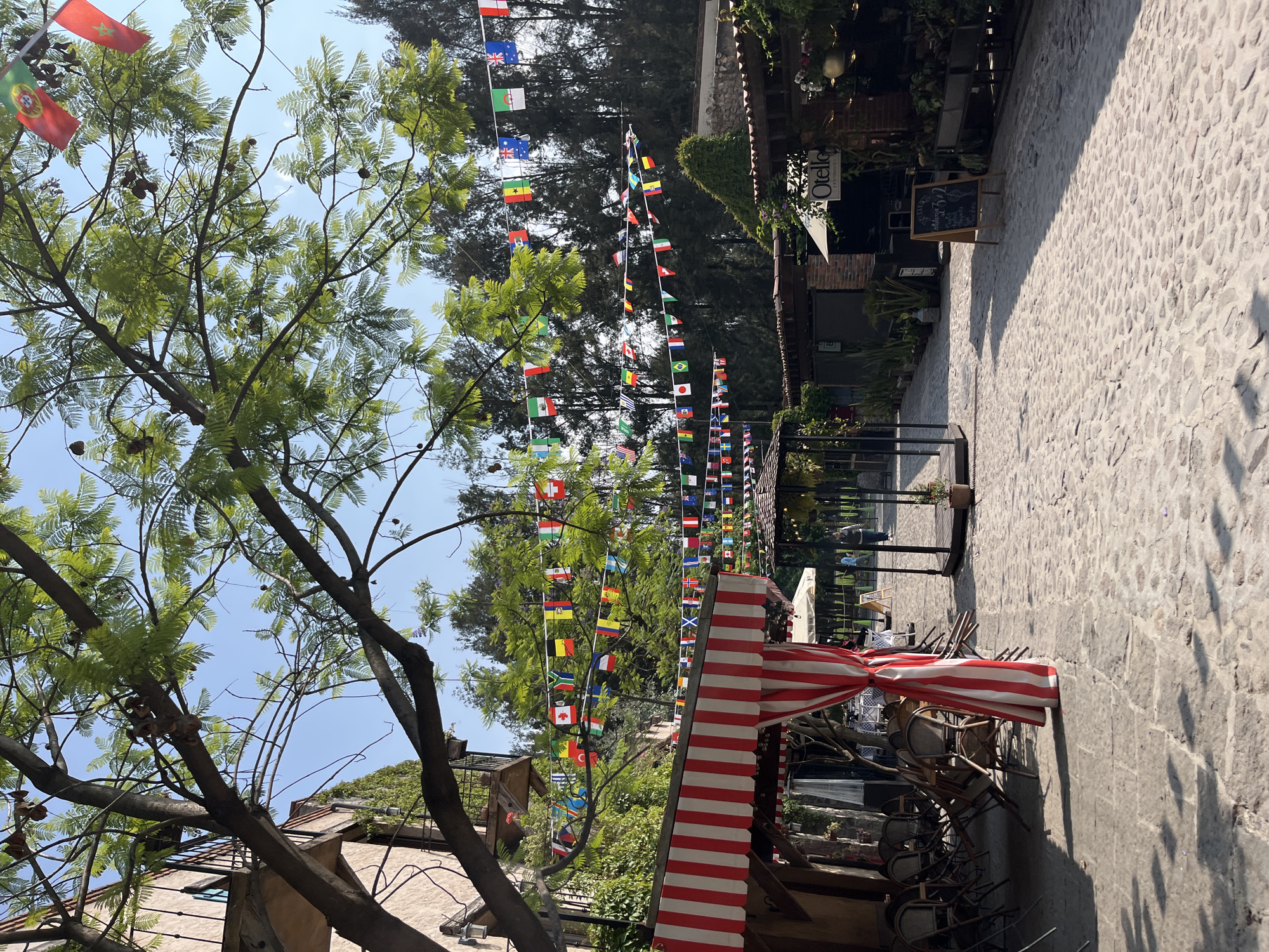 The image depicts a cobblestone street in what appears to be Domingo Arenas, Mexico. Strings of international flags are strung across the street overhead. A tree with small, light green leaves and dangling brown seed pods is visible to the left. Seating under a red and white striped awning is visible along the right side of the street. A building with a sign reading "Otello" is seen further down the street. The scene is sunlit. There are no people visible.
