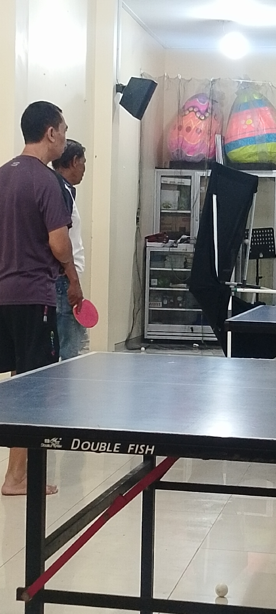 In Kudus, Indonesia, a ping pong table, branded "DOUBLE FISH," is positioned in a room with light-colored walls and tiled flooring. Two men are present. One man, on the left, is wearing a purple t-shirt and black shorts, his left hand holding a pink ping pong paddle. The other man, to his right, wears a white t-shirt with blue accents and jeans. He appears to be looking away from the paddle. In the background, a black speaker is mounted on the wall, and behind a screen, colorful, egg-shaped decorations are visible. A tall, white cabinet with glass doors, containing various items, stands to the right, partially obscured by a black fabric draped over a stand. A music stand is also visible near the cabinet. A white ping pong ball rests on the floor near the table.