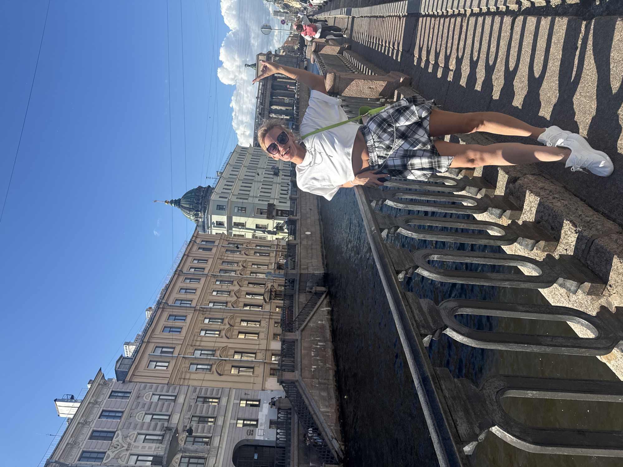 A young woman stands on a bridge in Saint Petersburg, Russia, her left hand resting on the ornate metal railing as she smiles and gives a rock-and-roll hand gesture with her right hand. She is wearing a white crop top, a black and white plaid skirt, sunglasses, and white sneakers. A bright green bag hangs from her left shoulder. Behind her, the Griboyedov Canal flows, lined with historic buildings. The canal is bordered by a stone embankment with a prominent stone staircase leading down to the water. The architecture visible includes classical buildings with tall windows and decorative facades, as well as a domed building in the distance. The sky is clear blue with scattered white clouds, and shadows from the railing and the woman are cast onto the paved walkway. Several other people are visible further down the bridge, walking away from the camera.