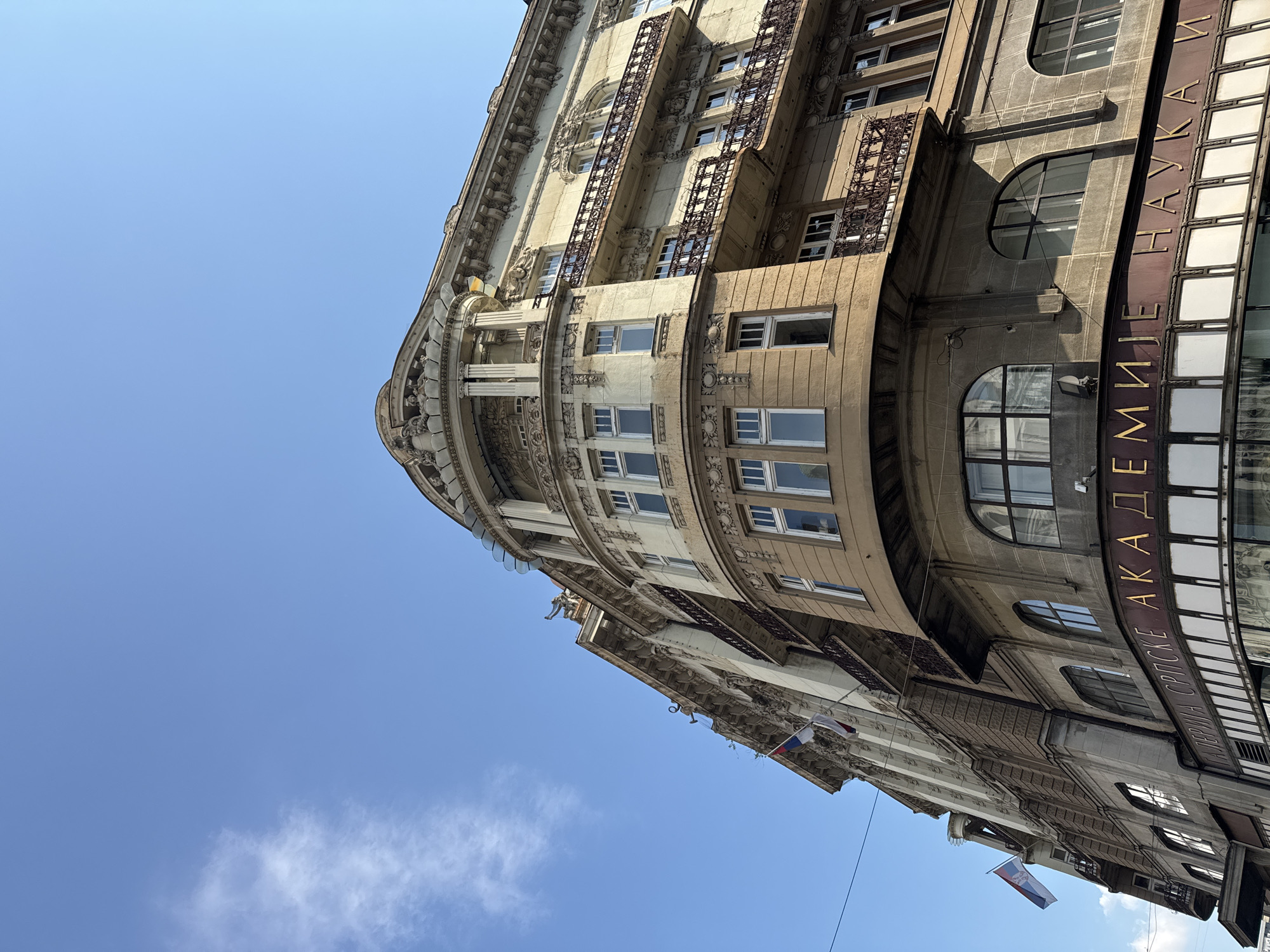 The image shows the Serbian Academy of Sciences and Arts building, located in Belgrade, Serbia. The building is captured from a low angle, emphasizing its ornate architecture and the bright blue sky above. The facade features classical detailing, including columns, balconies with decorative ironwork, and arched windows. Several Serbian flags are visible on the building. Below the main facade, a sign with Cyrillic lettering, "СРПСКЕ АКАДЕМИЈЕ НАУКА И", is prominently displayed. The image is taken during the day, with clear weather and a few wispy clouds. There are no people or vehicles visible in the frame.