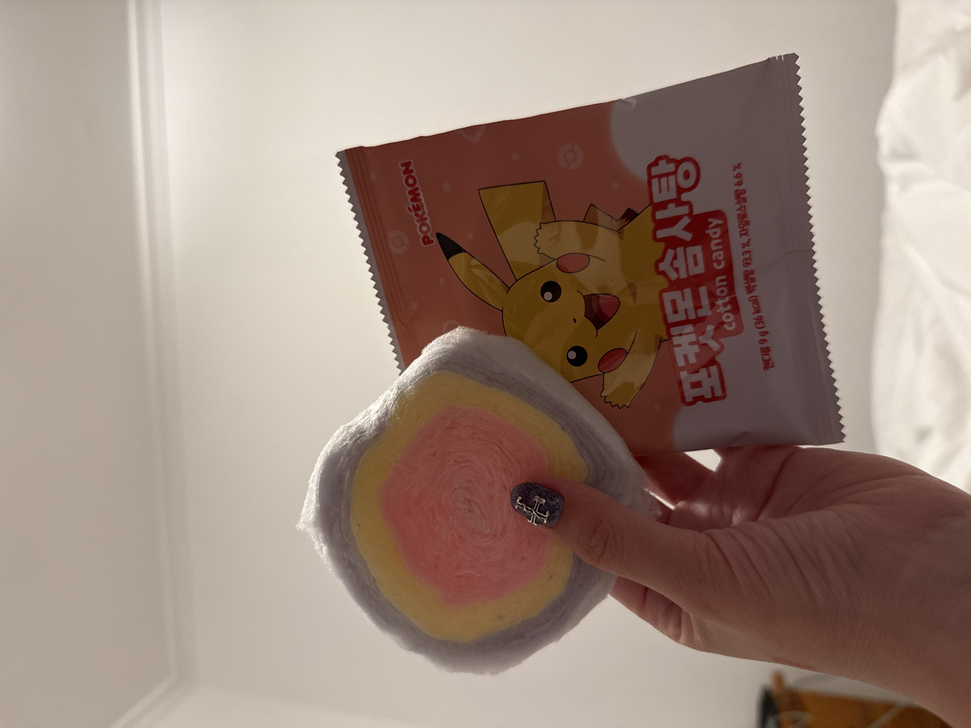 The image shows a person's hand holding a piece of cotton candy and a package of Pokémon cotton candy. The cotton candy is round and fluffy, with layers of gray, yellow, and pink. The package is pink and white, with an image of Pikachu on it. The background is a white wall.
