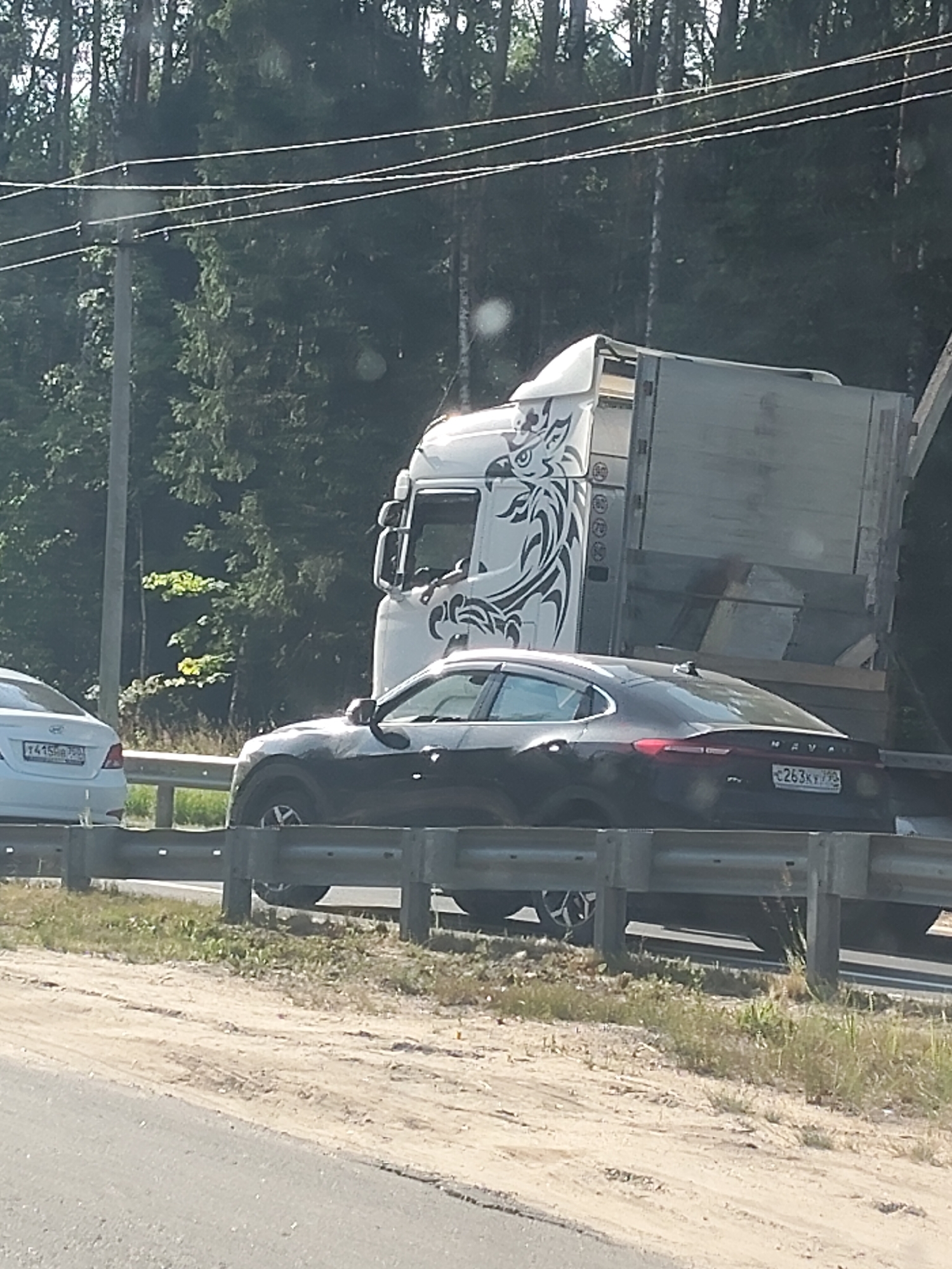 In this image, a dark-colored sedan is on a highway, partially obscured by a large white truck. The sedan's rear is visible, displaying a license plate that reads "E263KY 790". The truck has a distinctive tribal-like graphic on its side, resembling a stylized bird or animal. The scene appears to be on a sunny day, with lush green trees lining the background. A guardrail separates the highway from a grassy and sandy shoulder. Another white sedan is partially visible in the far left of the image, with its license plate showing "T415CB 790". The overall setting suggests a rural or highway environment in Russia, possibly during daylight hours given the bright illumination and shadows. No specific activity or event is depicted, other than vehicles traveling on a road. The image is taken from a slightly elevated perspective, looking down at the vehicles and the road.