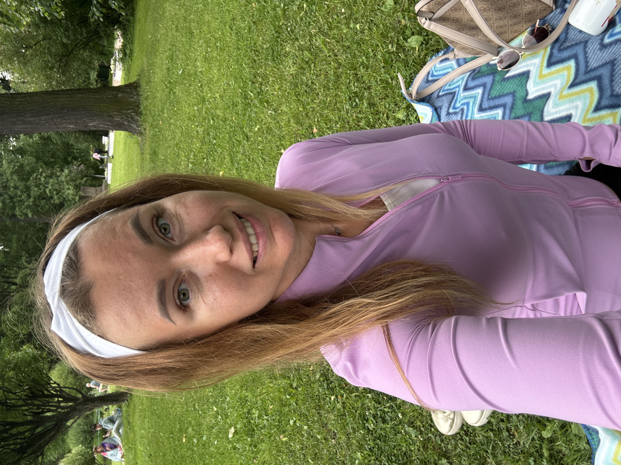 A woman with blonde hair and a white headband is taking a selfie in a park. She is wearing a light purple zip-up jacket over a white top. Her eyes are green, and she is smiling. Behind her, a picnic blanket with a zigzag pattern is spread on the grass, with a purse and sunglasses on it. Two pairs of white sneakers are visible near the blanket. In the background, other people are sitting on the grass and walking among trees. The environment is a grassy park with mature trees and lush green foliage.