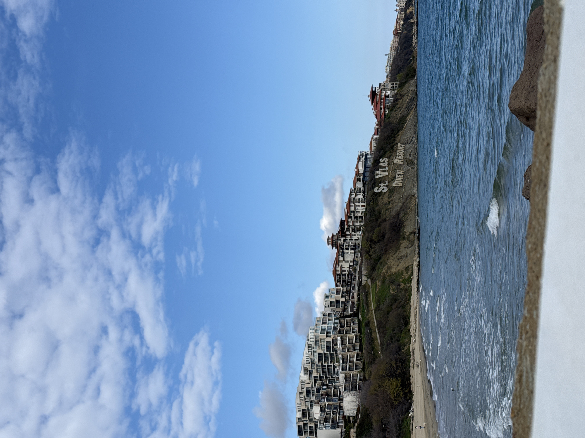 The image depicts a view of Sveti Vlas, Bulgaria, from an elevated position, possibly a balcony or ledge, suggested by the stone structure on the right side of the image. The sky is bright blue with scattered white clouds. The sea occupies the right side of the frame. The coastline slopes upwards, featuring buildings and vegetation. A large sign that reads "St. Vlas Dinevi Resort" is visible on the hillside. A sandy beach is visible at the base of the slope. Small figures of people are present on the beach, but are too small to analyze.