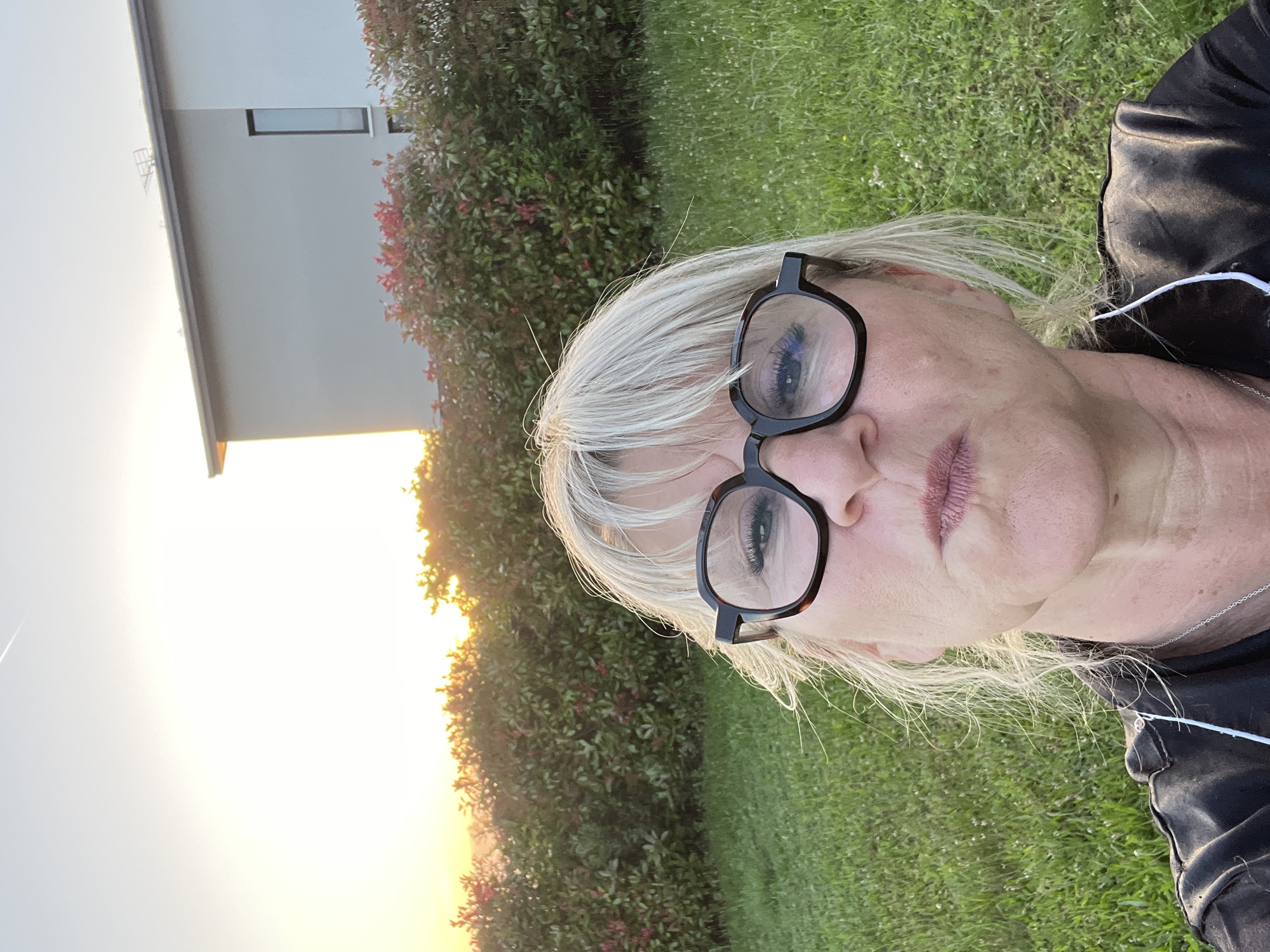 A woman wearing black-rimmed glasses and a black jacket is facing the camera, in a landscape setting. She has short, gray hair and is wearing earbuds. Behind her is a building with a light-colored facade and a single visible window. A dense hedge with reddish leaves is situated between the woman and the building. The setting sun is visible on the horizon, casting a warm light on the scene. The ground is covered in green grass. The location is Chambœuf, France.