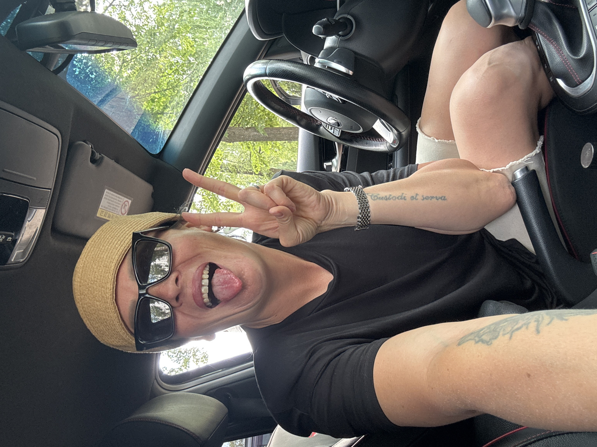 A woman wearing a straw hat and sunglasses is in the driver's seat of a car. She has her tongue sticking out and is making a peace sign with her fingers. The car is parked outdoors, with trees and foliage visible through the windows. The woman has tattoos on her arm. The interior of the car includes a steering wheel, gear shift, and center console. There is no indication of the city of Chaikovsky, Russia, in the image.